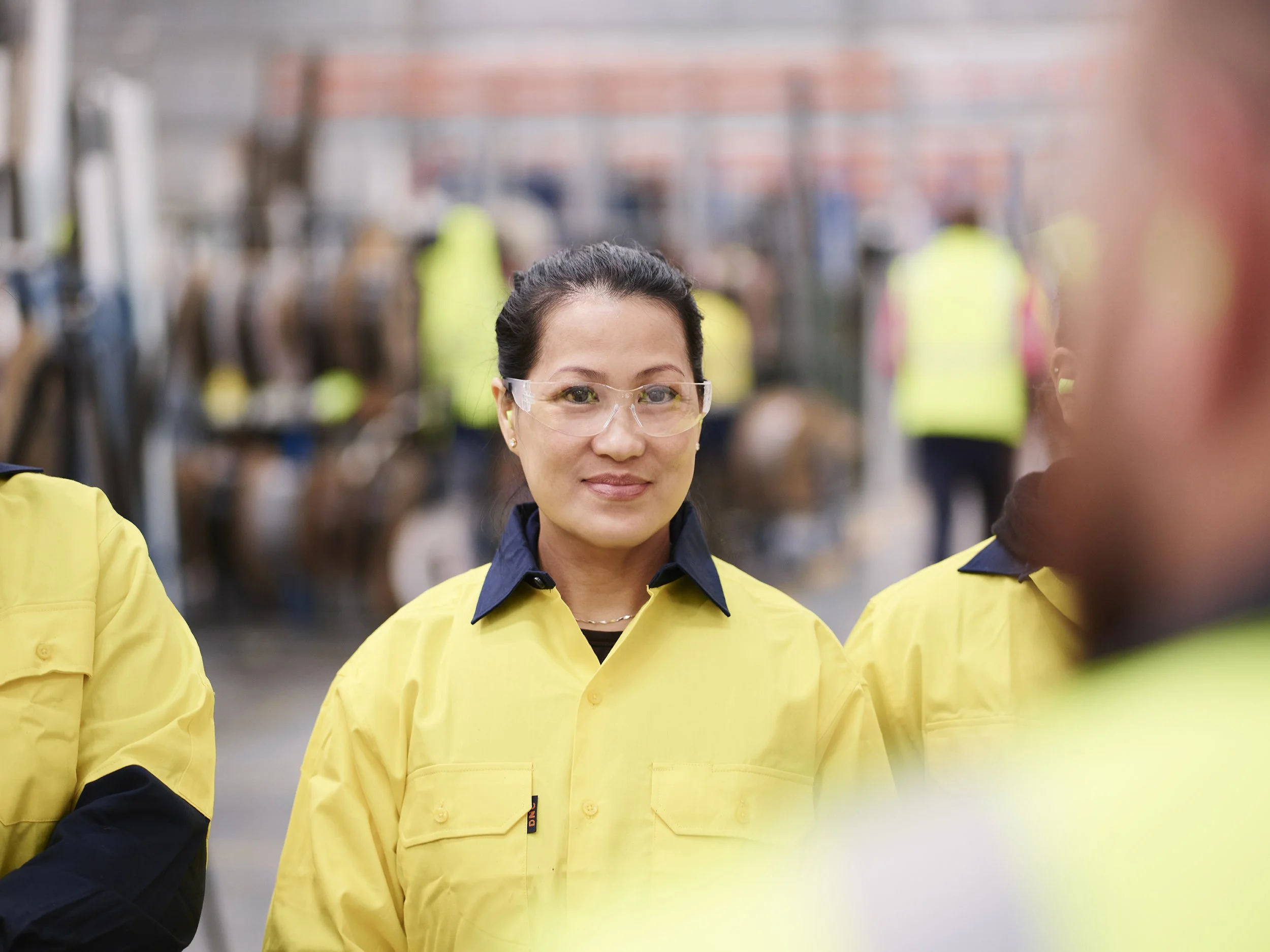 A woman wearing safety glasses and a yellow safety jacket with a dark collar, smiling at the camera. She is outdoors with a blurred background of other people in similar safety gear and various equipment.