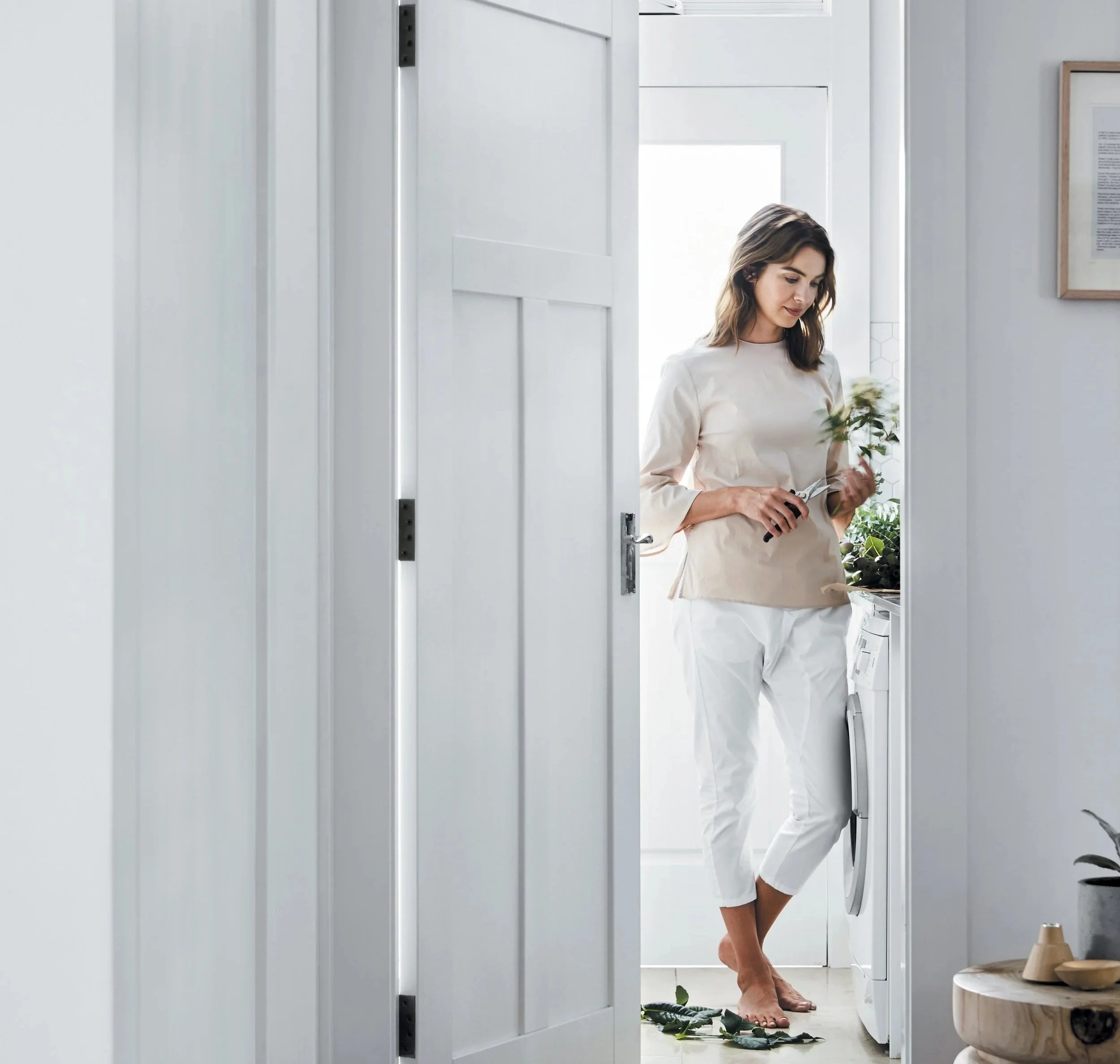 A woman with shoulder-length brown hair standing in a laundry room, holding a plant and a pair of scissors, barefoot, near a washing machine and plants, with a white door and a framed document on the wall.