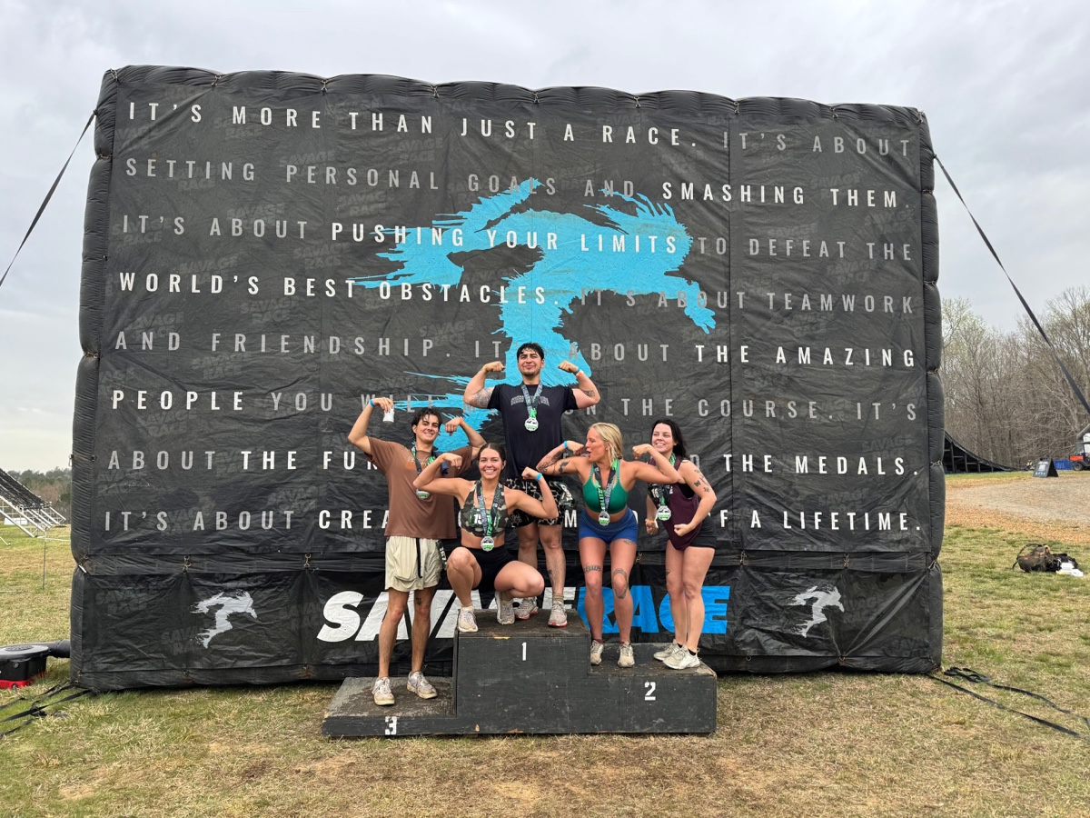 Group of five runners on a winners' podium celebrating after completing a race, with medals around their necks, in front of a large black banner with motivational text and a blue graphic.