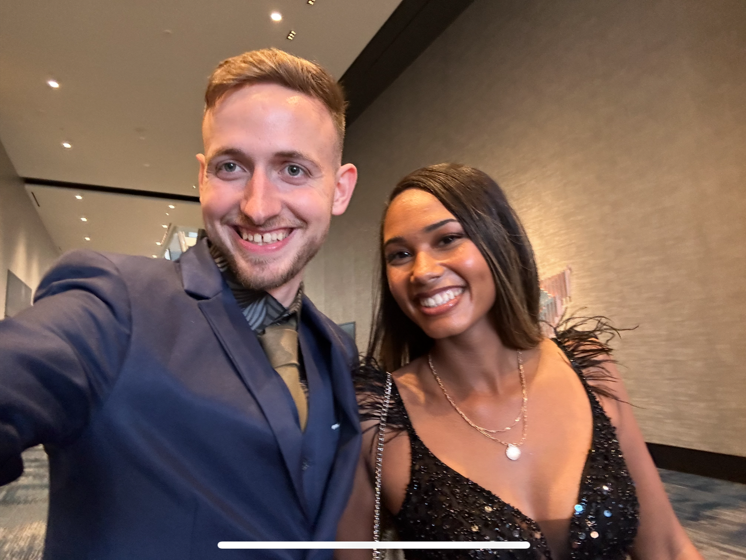 A young man and woman smiling for a selfie at an indoor event. The man has short blond hair, a beard, and is wearing a navy blazer over a dark shirt with a patterned tie. The woman has long dark hair, a bright smile, and is dressed in a black, sequin