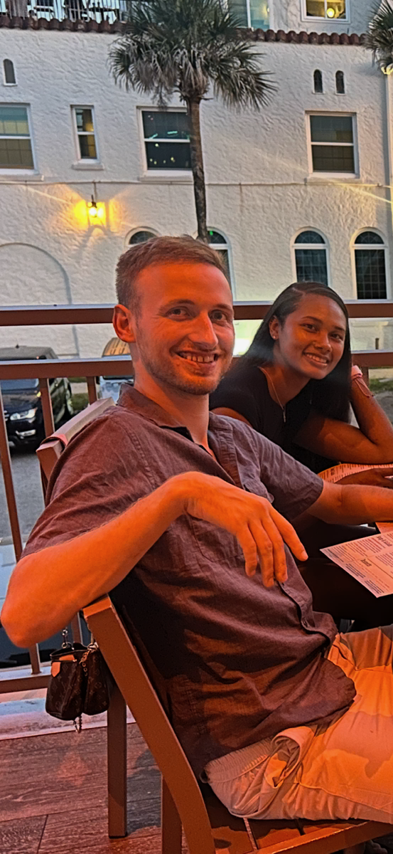 A smiling man and woman sitting at an outdoor restaurant table during sunset, with a white building, balcony, and palm tree in the background.
