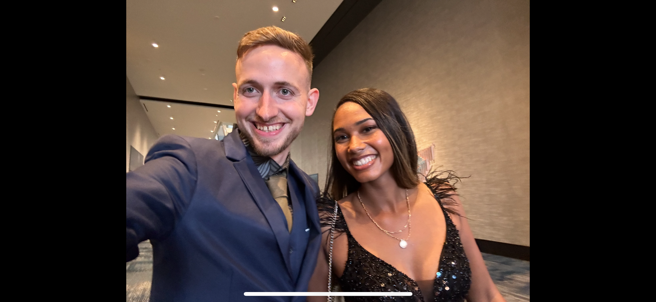 A man and woman dressed elegantly, smiling at the camera, in a well-lit indoor setting.