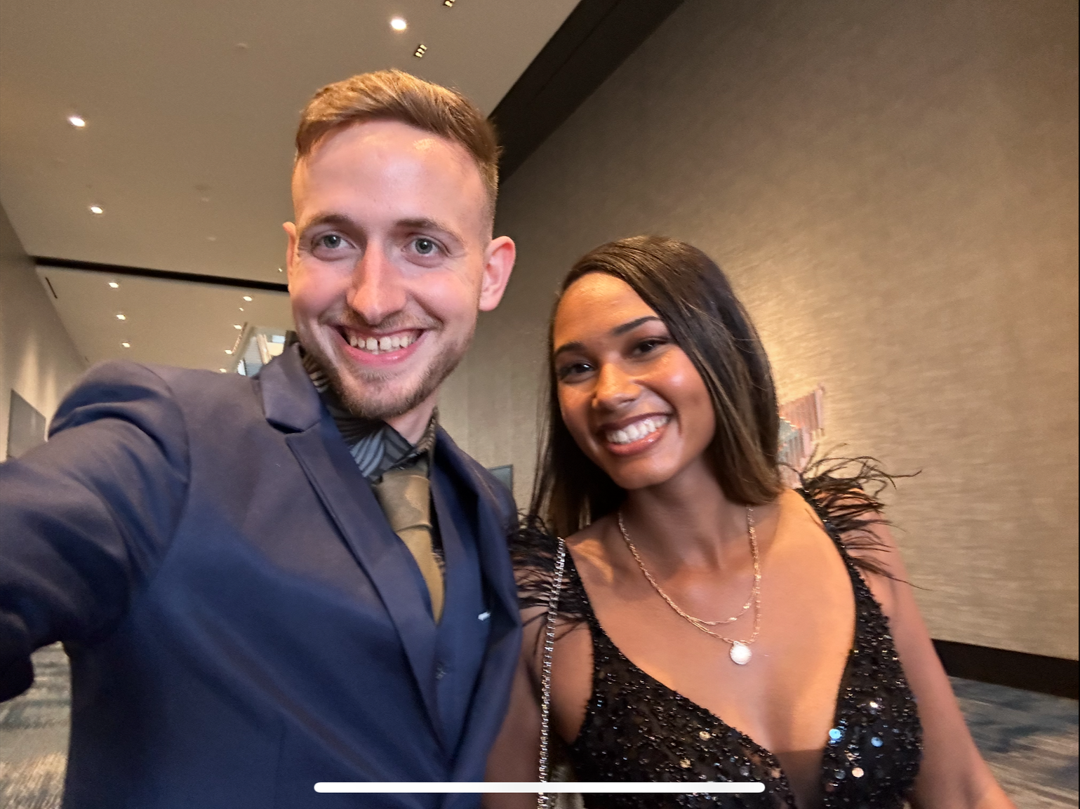 A man and woman taking a selfie at an indoor event, both smiling, wearing formal attire. The man has short hair and a beard, dressed in a dark suit with a striped shirt and tie. The woman has long dark hair, wearing a black sequined dress and layered gold necklaces.