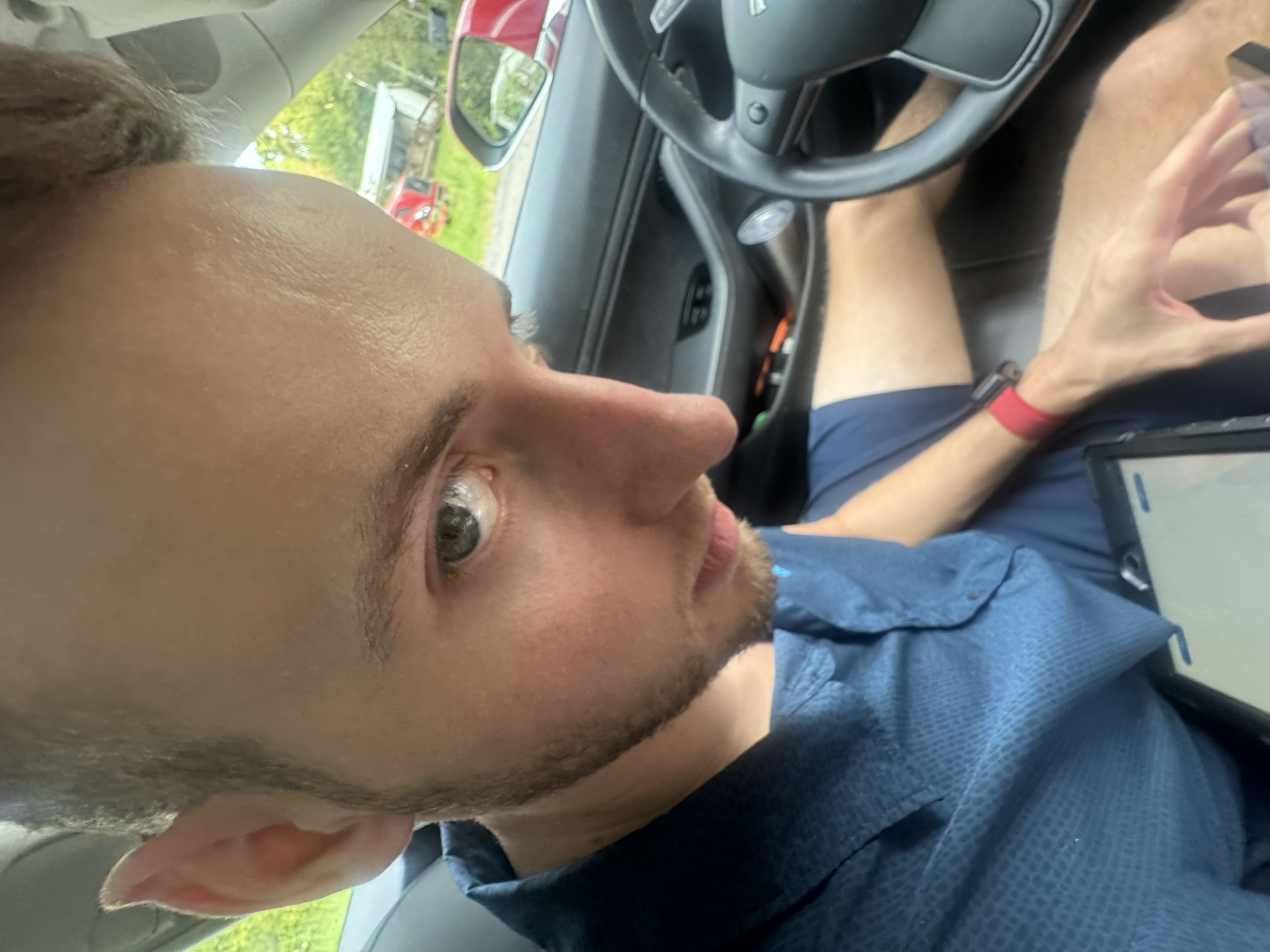 A man with a beard and short hair sitting in the driver's seat of a car, looking up at the camera, with a phone or tablet in his hand and an outdoor scene visible through the window.