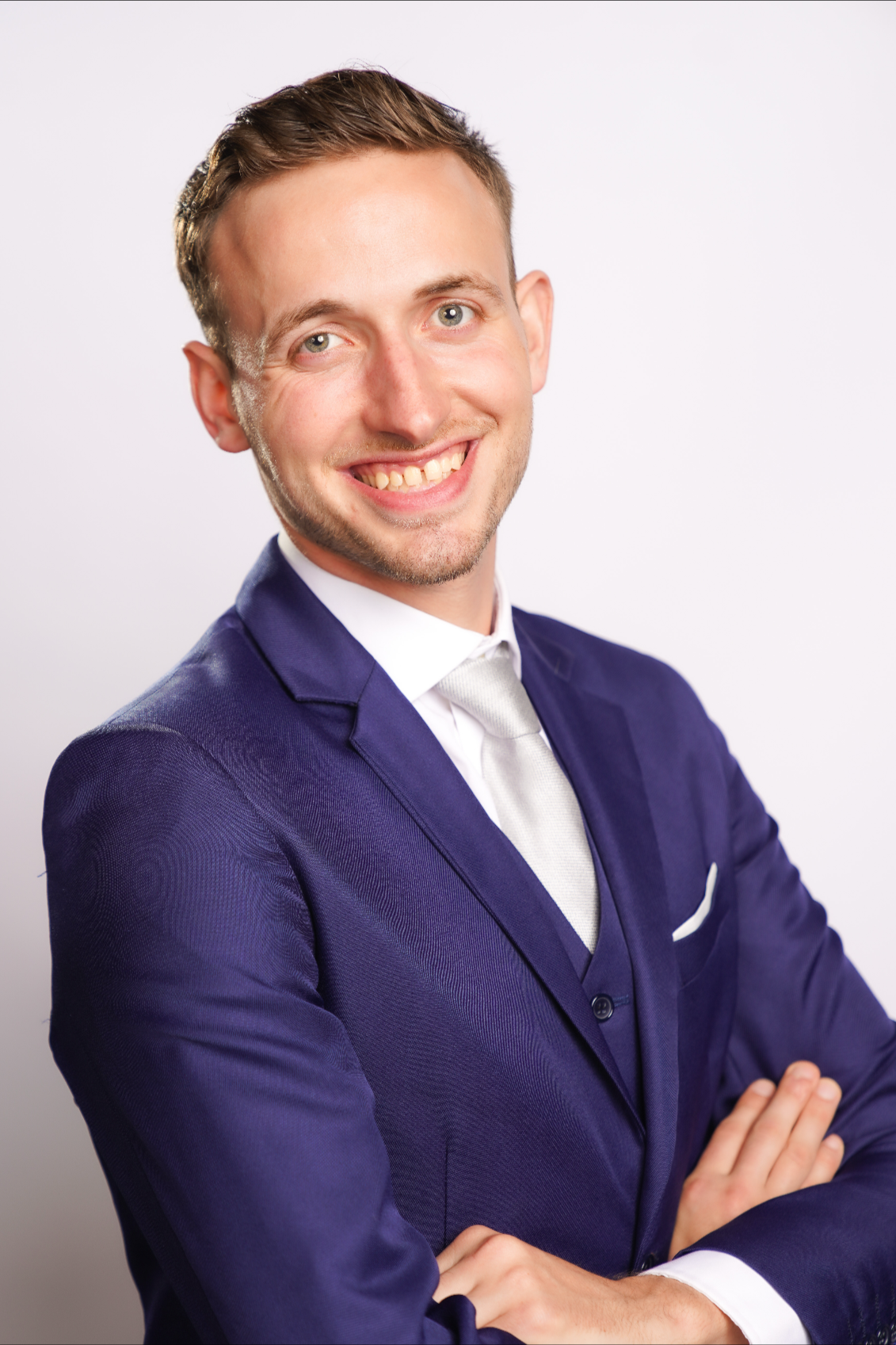 A smiling young man with light skin, wearing a navy blue suit, white shirt, and silver tie, standing with arms crossed against a plain white background.