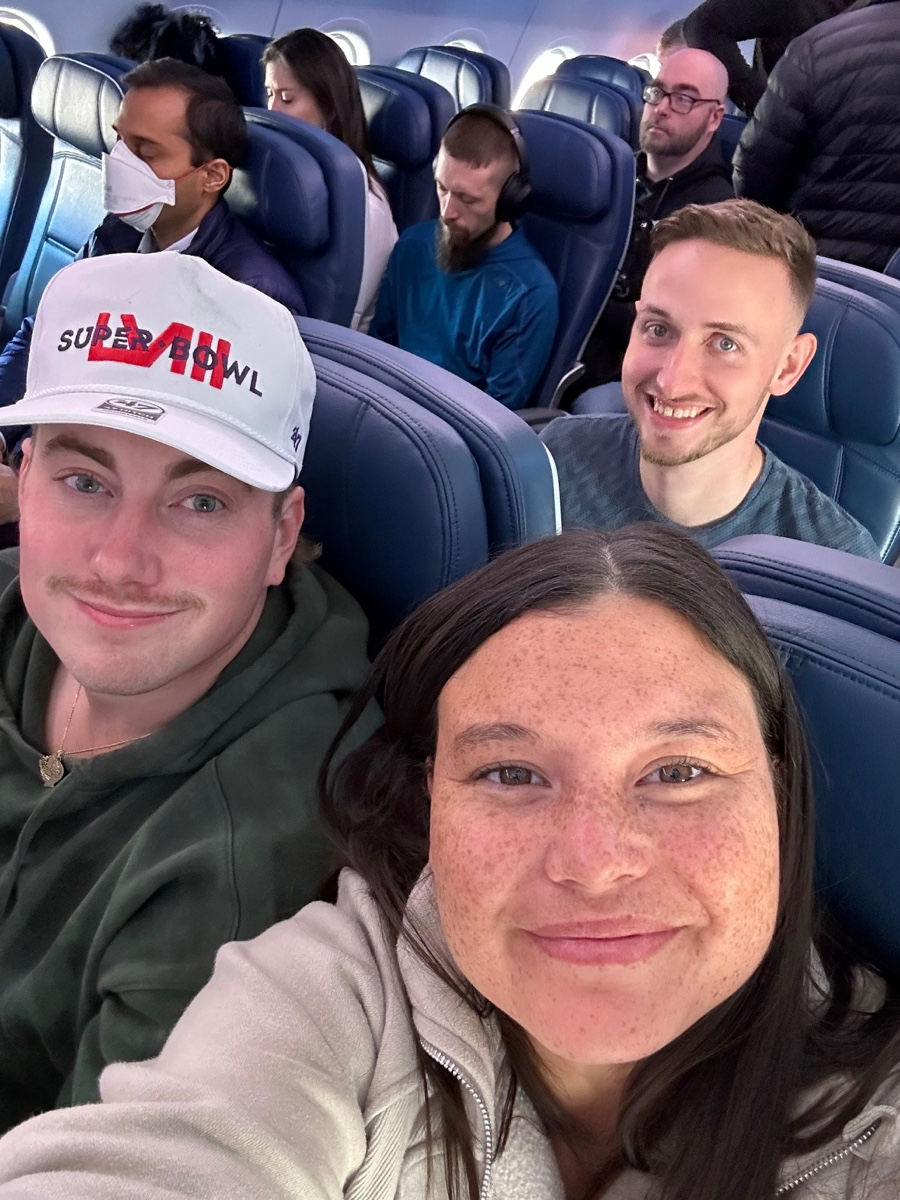 Group of five people sitting on an airplane, taking a selfie. Two women and three men, some wearing headphones and face masks, smiling or appearing relaxed.