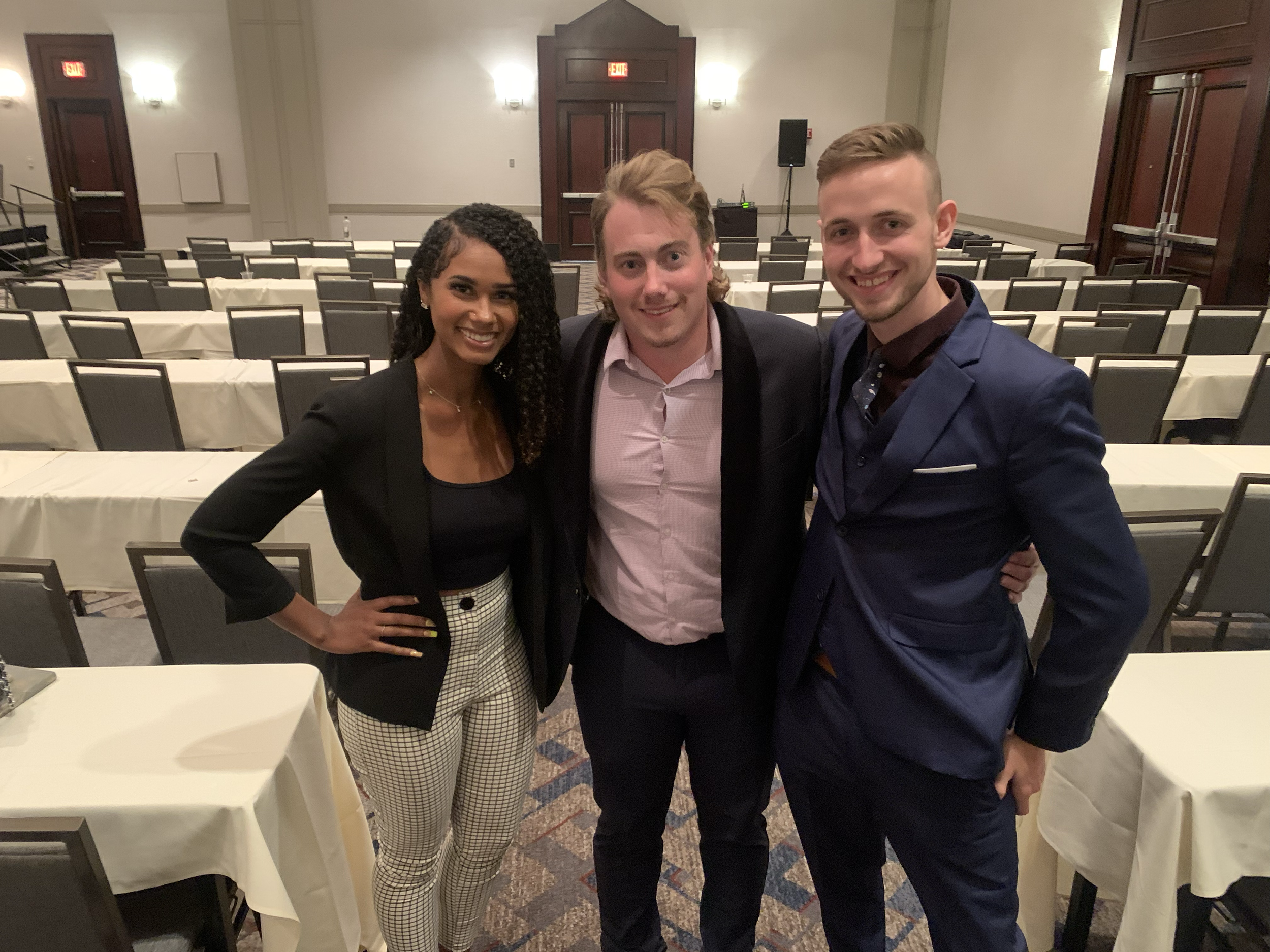 Three smiling young adults, two men and one woman, standing together in a conference or banquet room with rows of tables and chairs in the background.