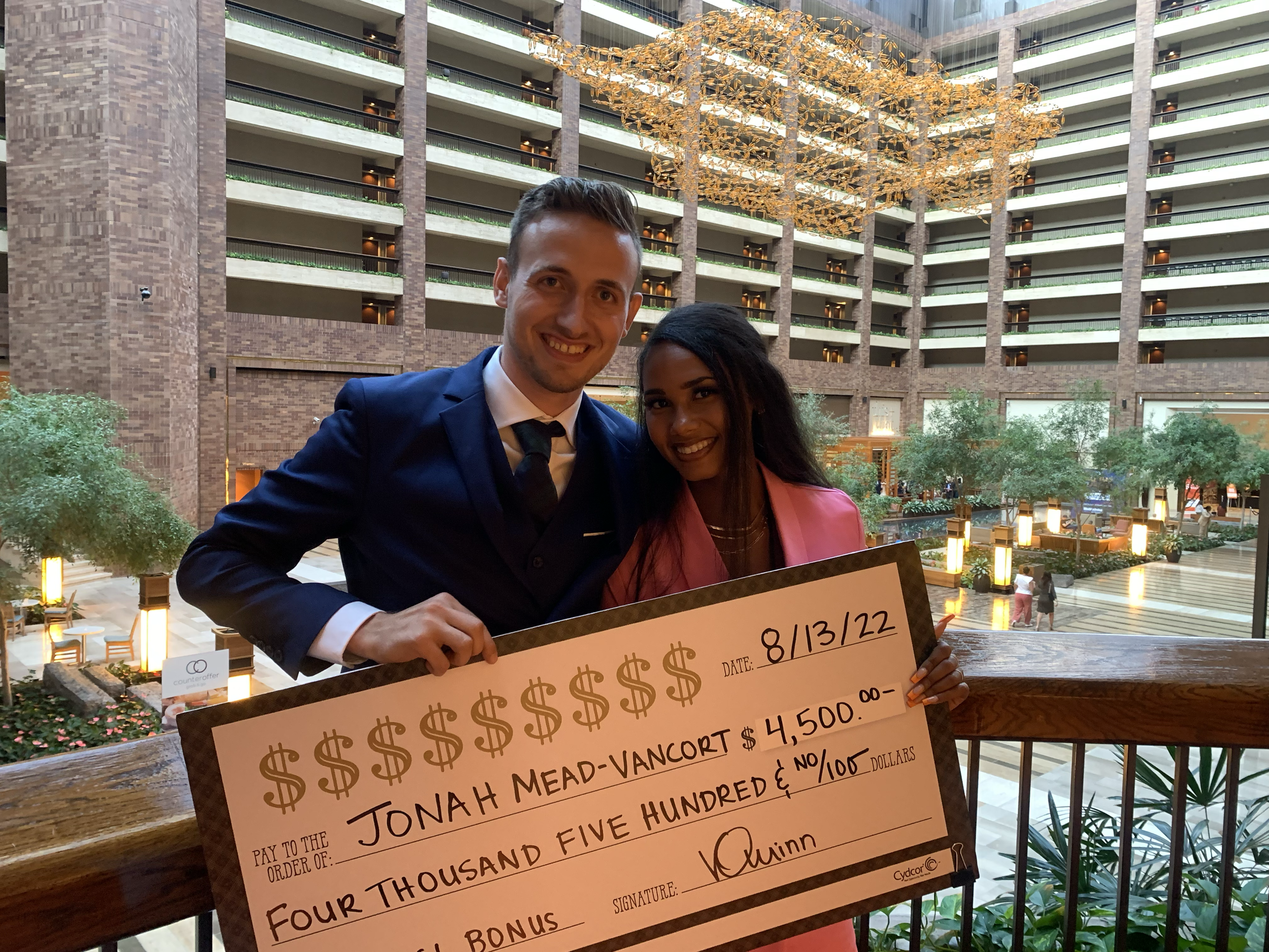 A man in a blue suit and a woman in a pink blazer holding a giant check for $4,500 at an indoor hotel or conference center with a glass ceiling and a courtyard below.