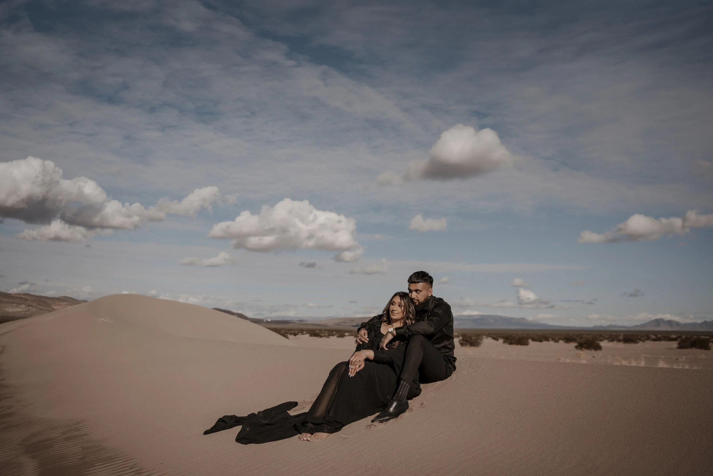 A couple dressed in black clothing sitting and lying on sand dunes in a desert landscape with a cloudy sky.