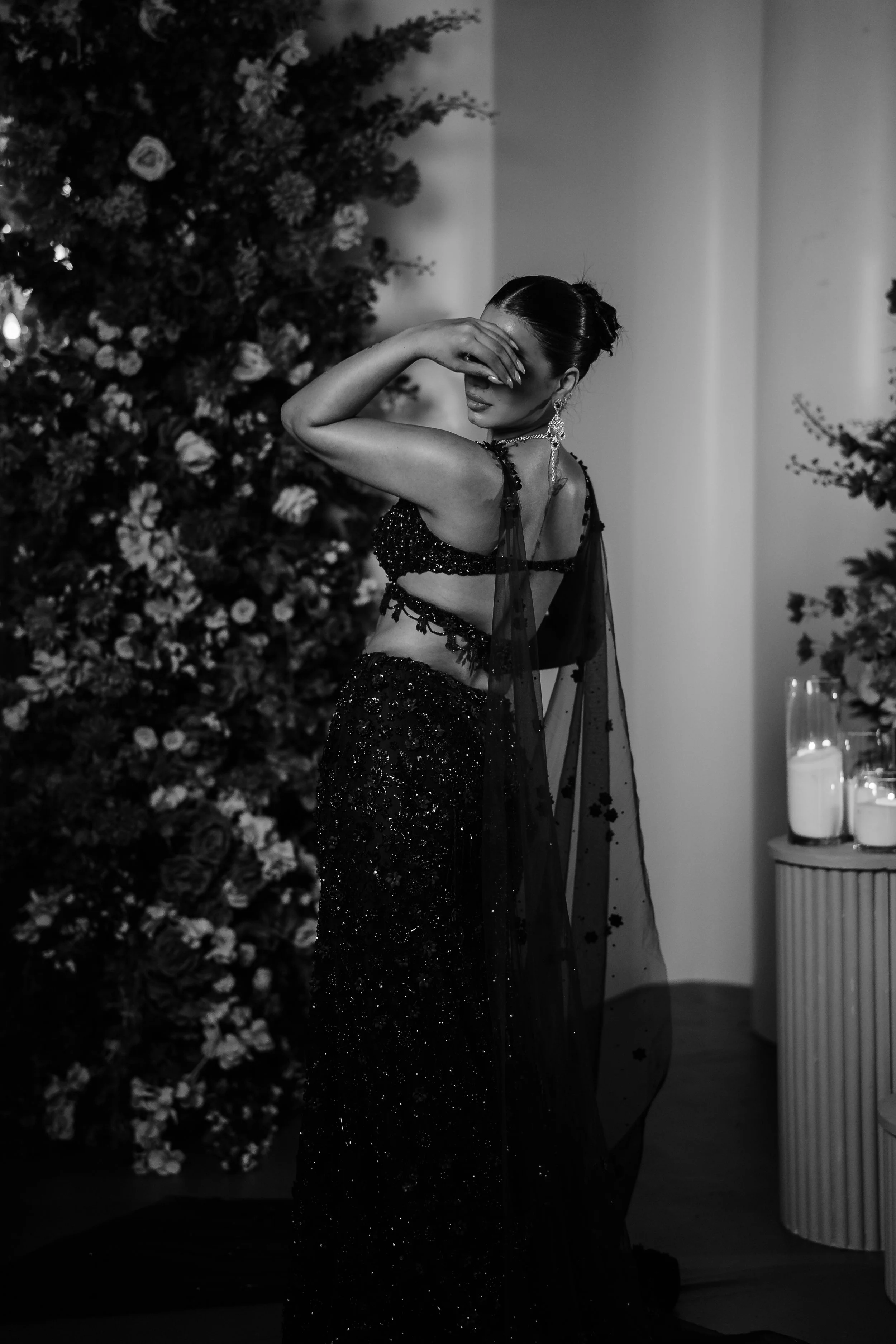 A woman in a sequined black evening gown featuring a sheer shawl, standing in front of floral decor, covering her eyes with one hand, in a monochrome setting.