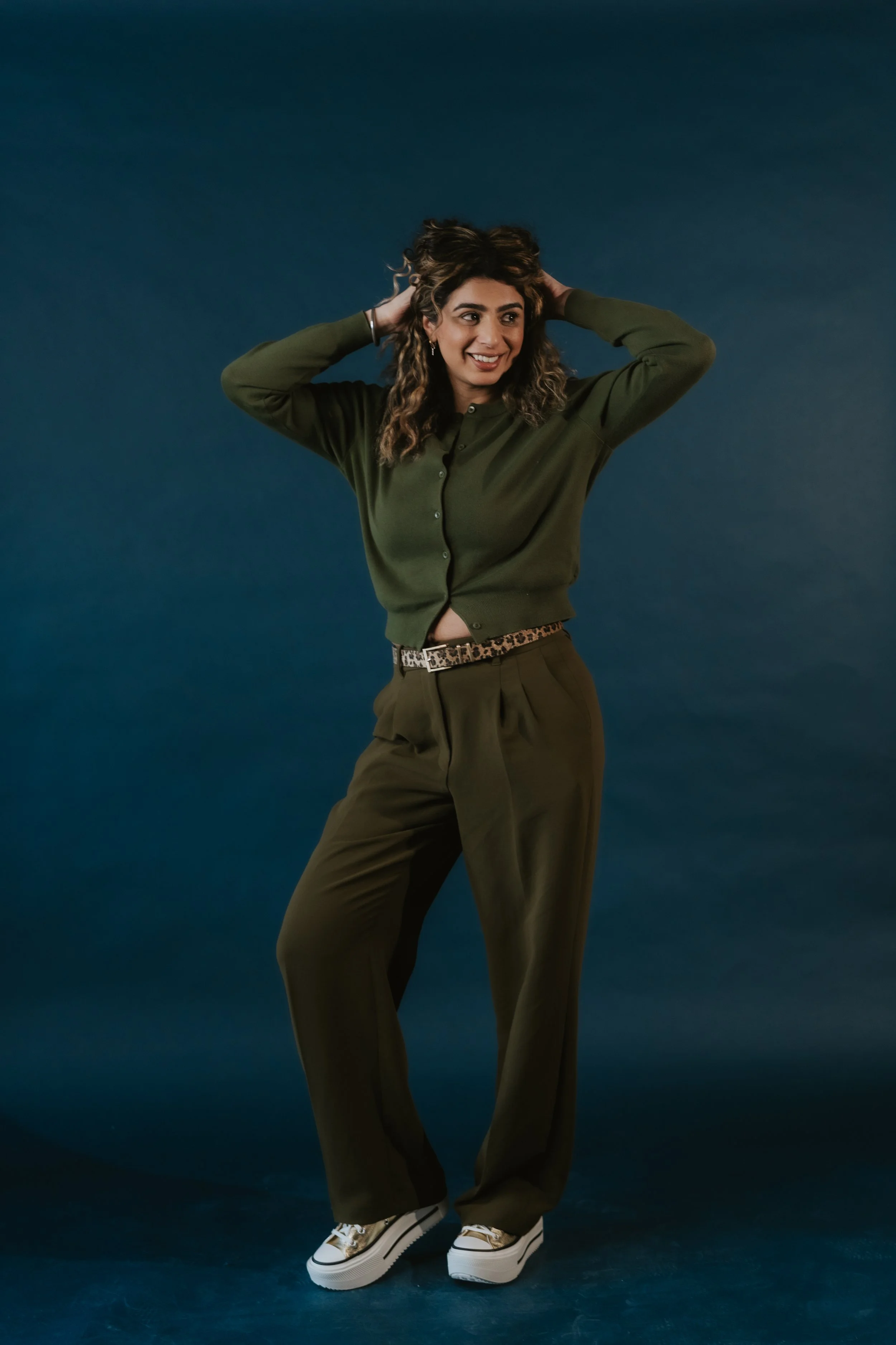 Young woman in a dark green cardigan and matching wide-legged pants, standing against a dark blue studio background, smiling with her hands in her hair, wearing white platform sneakers and a leopard print belt.