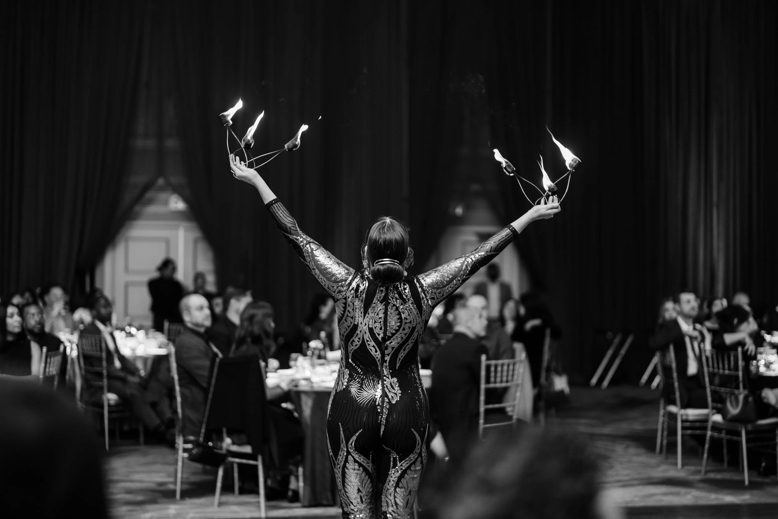 A performer in a patterned dress twirling fire poi in front of a seated audience at an indoor event.