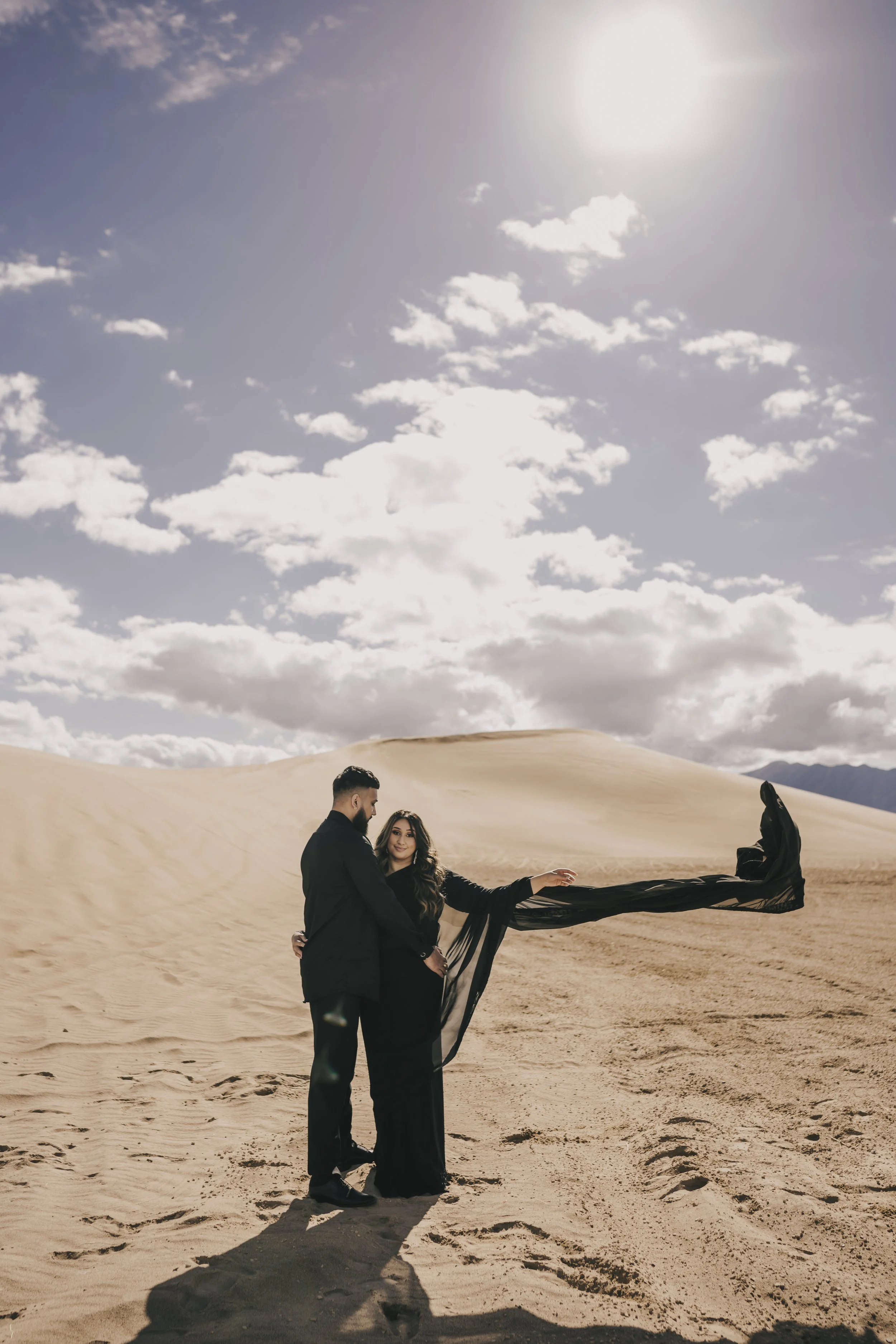 A couple dressed in black standing on sand dunes under a partly cloudy sky with the sun shining.