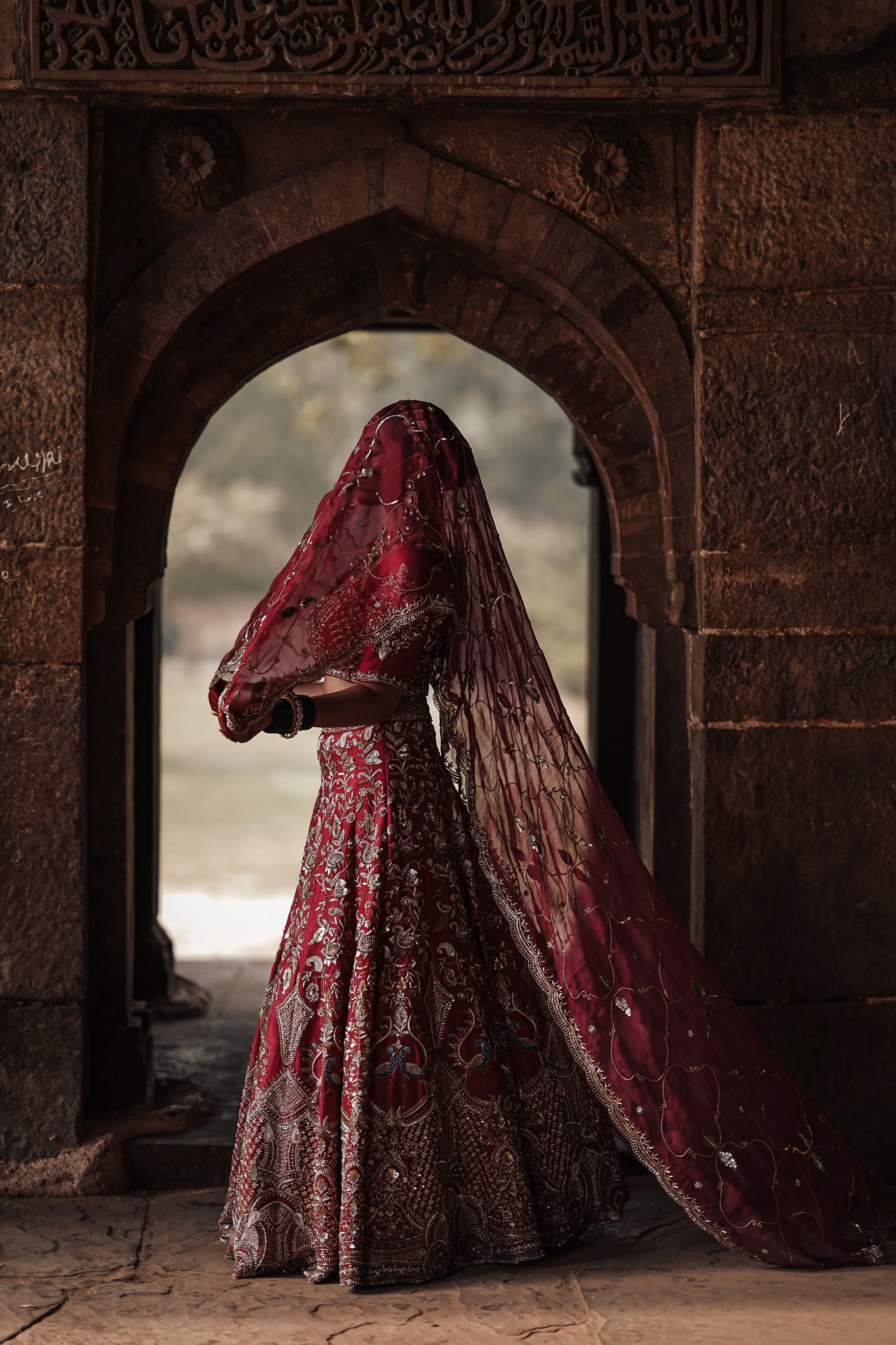 A woman dressed in a red and silver embroidered traditional Indian bridal outfit, standing under an arched stone doorway with intricate carvings, with her face partially covered by a red sheer veil.