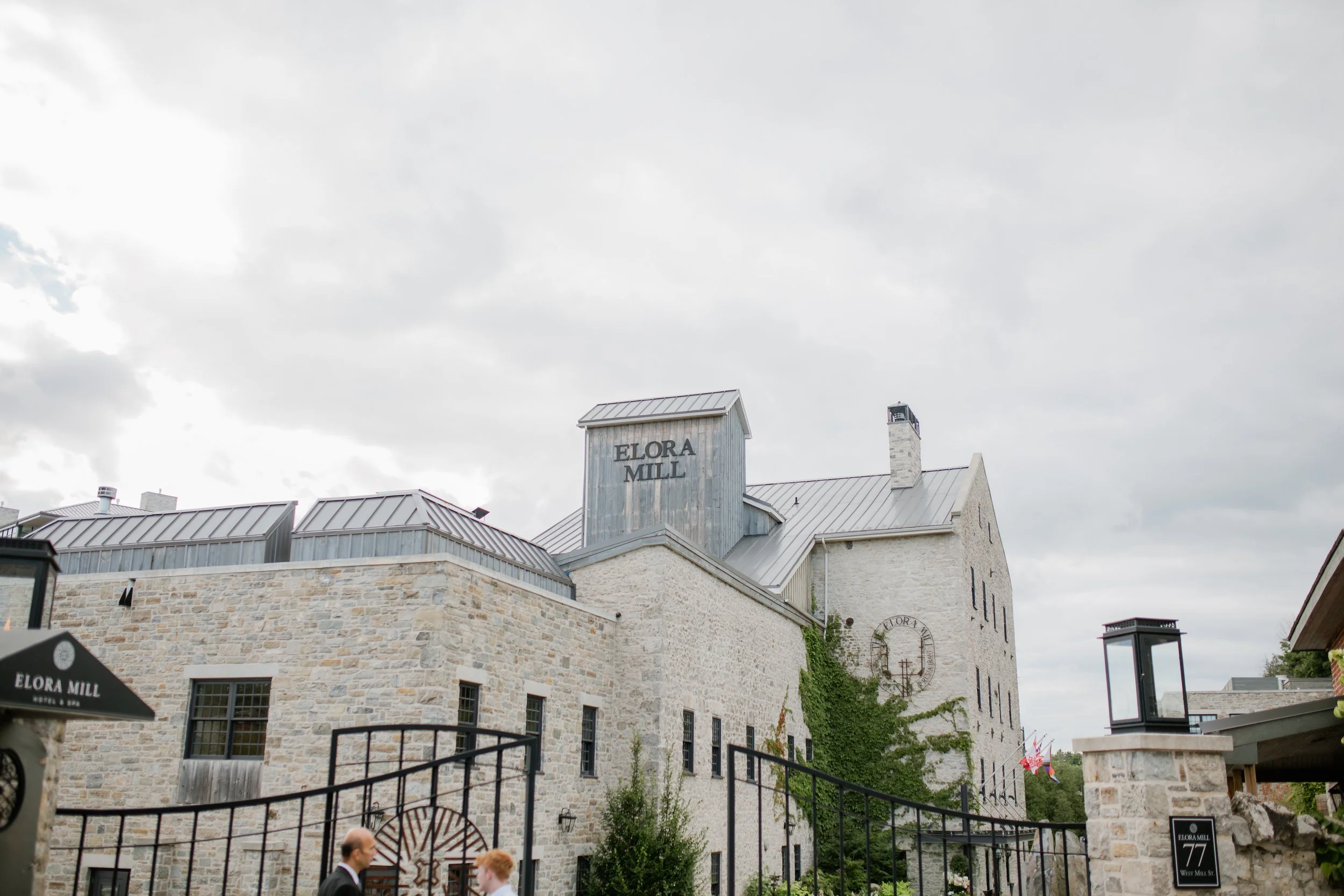 Exterior view of Elora Mill, a historic stone building with a metal roof, ivy growing on the walls, and signage displaying the name. There are people walking near the entrance, black metal gate fencing, and modern light fixtures.