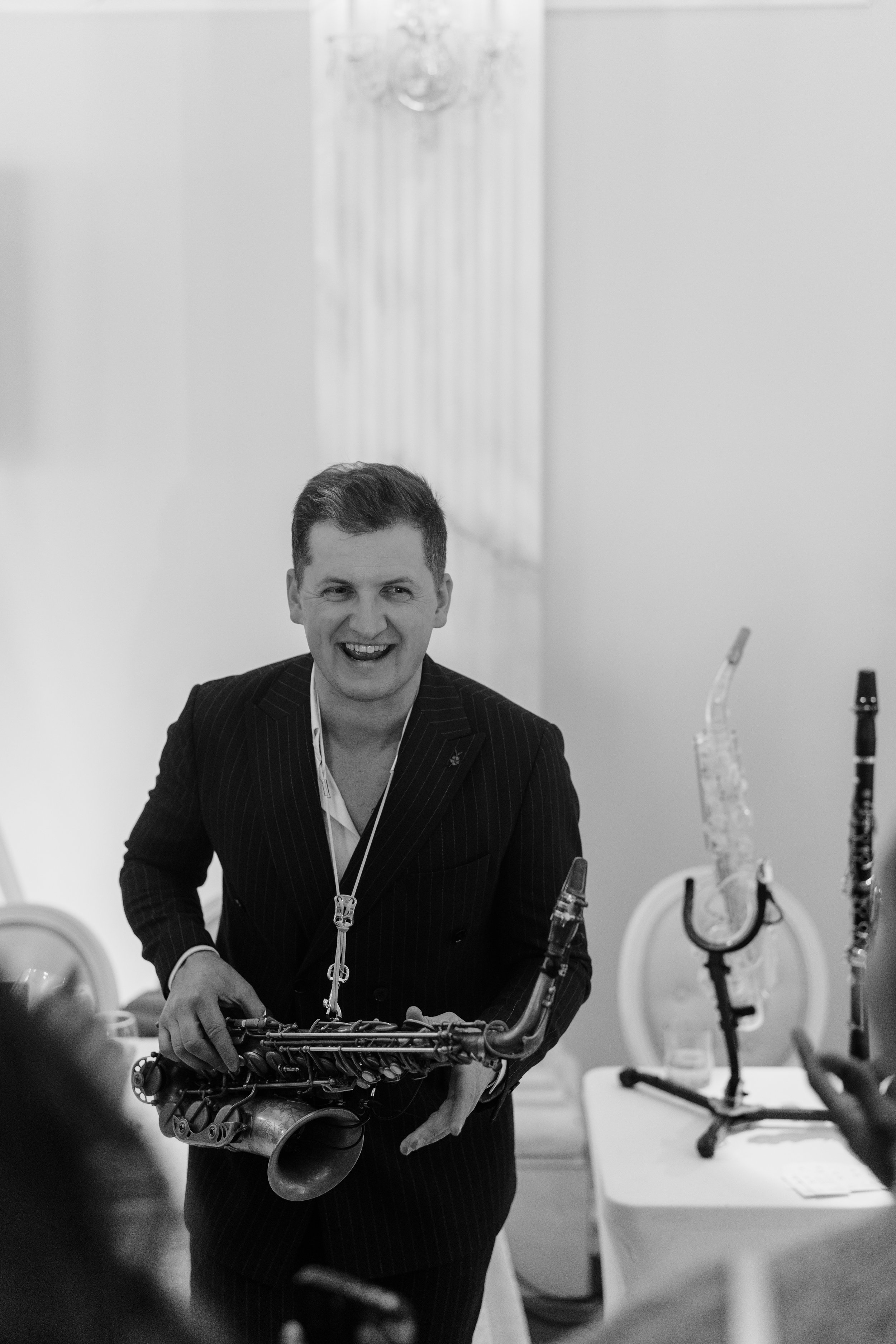 A man in a dark pinstripe suit holding a saxophone, smiling and interacting with others at a social event, with more musical instruments on display.