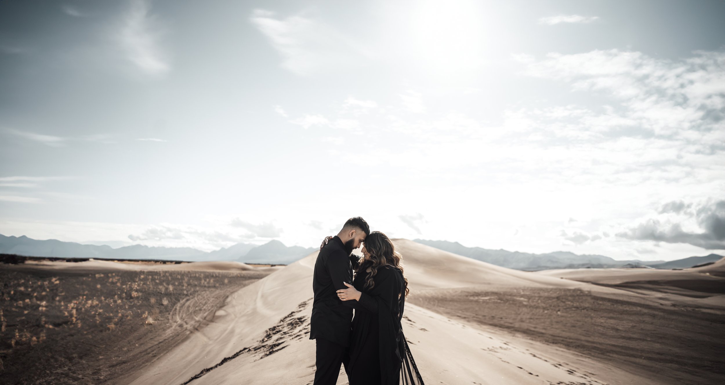 A couple embracing in a desert landscape with sand dunes and a cloudy sky in the background.