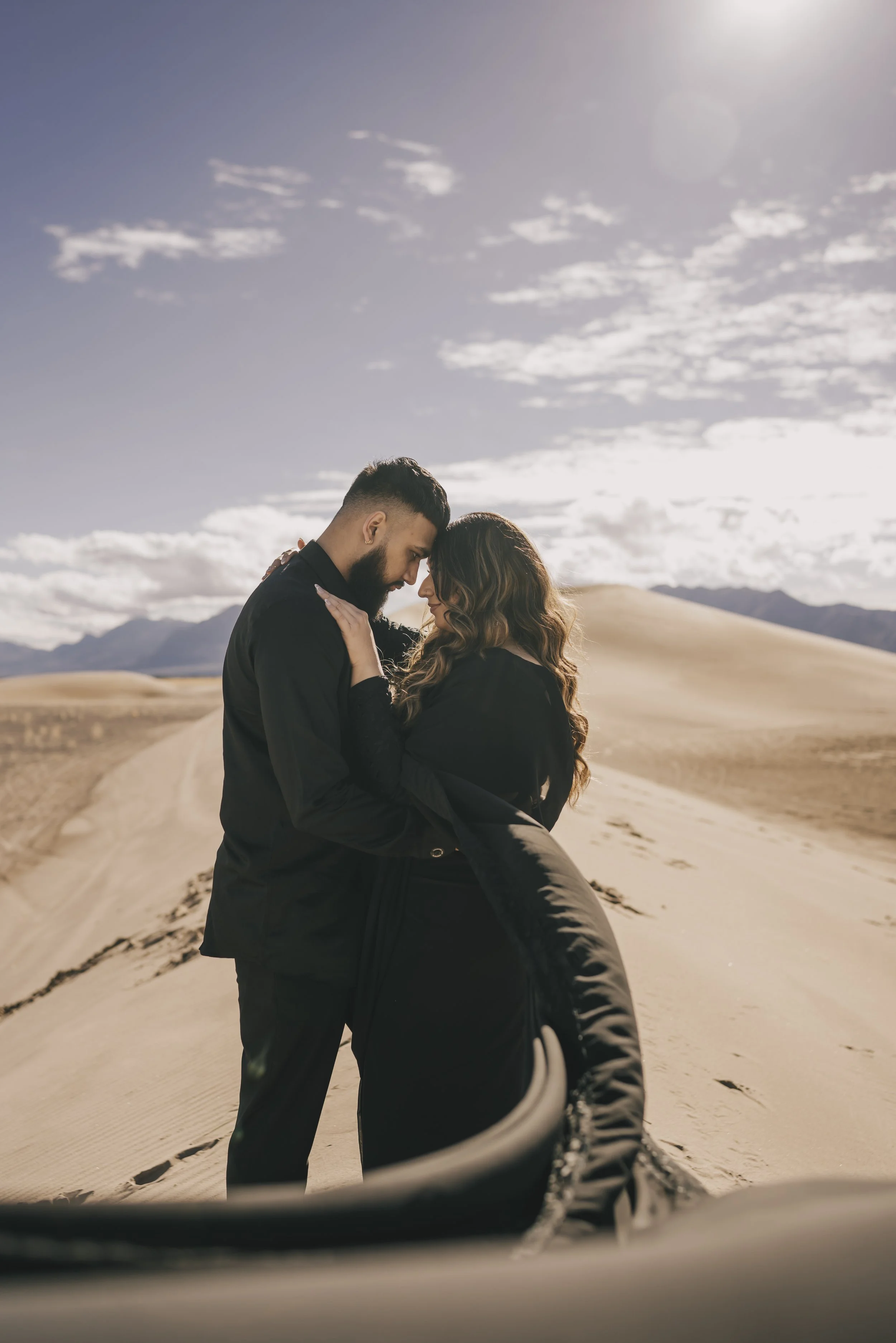 A couple dressed in black embracing in a desert landscape with sand dunes and mountains in the background under a partly cloudy sky.