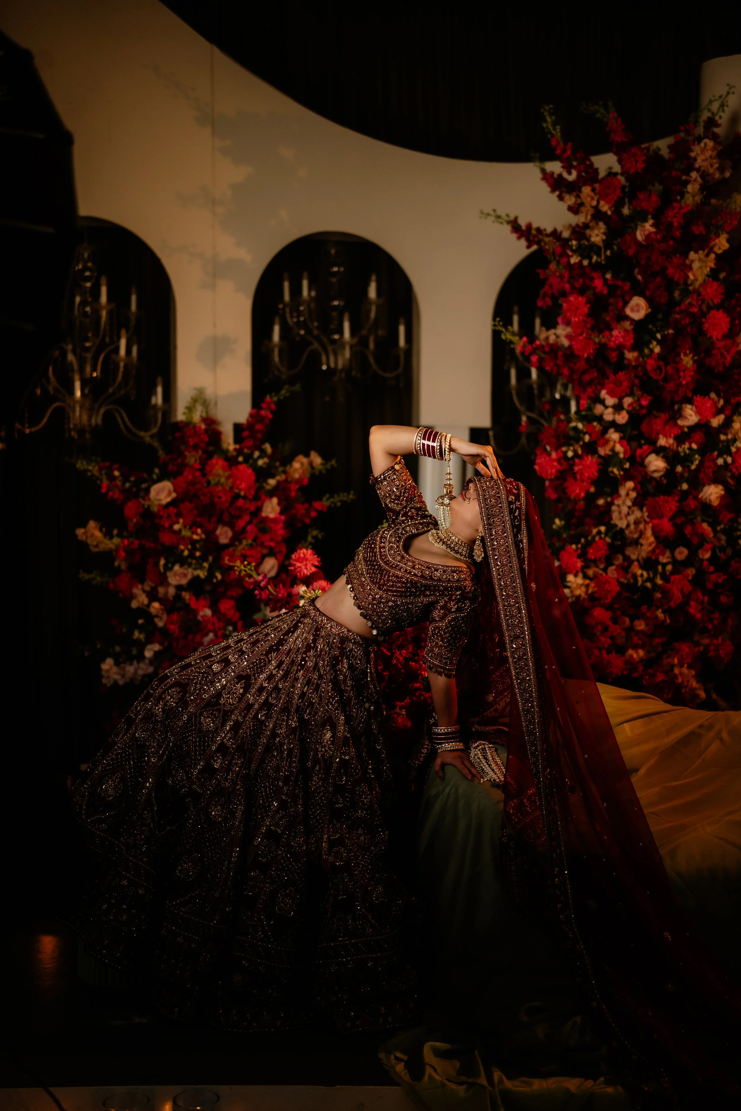 A woman dressed in traditional Indian bridal attire, including a richly embroidered lehenga and veil, poses in front of floral arrangements and chandeliers.