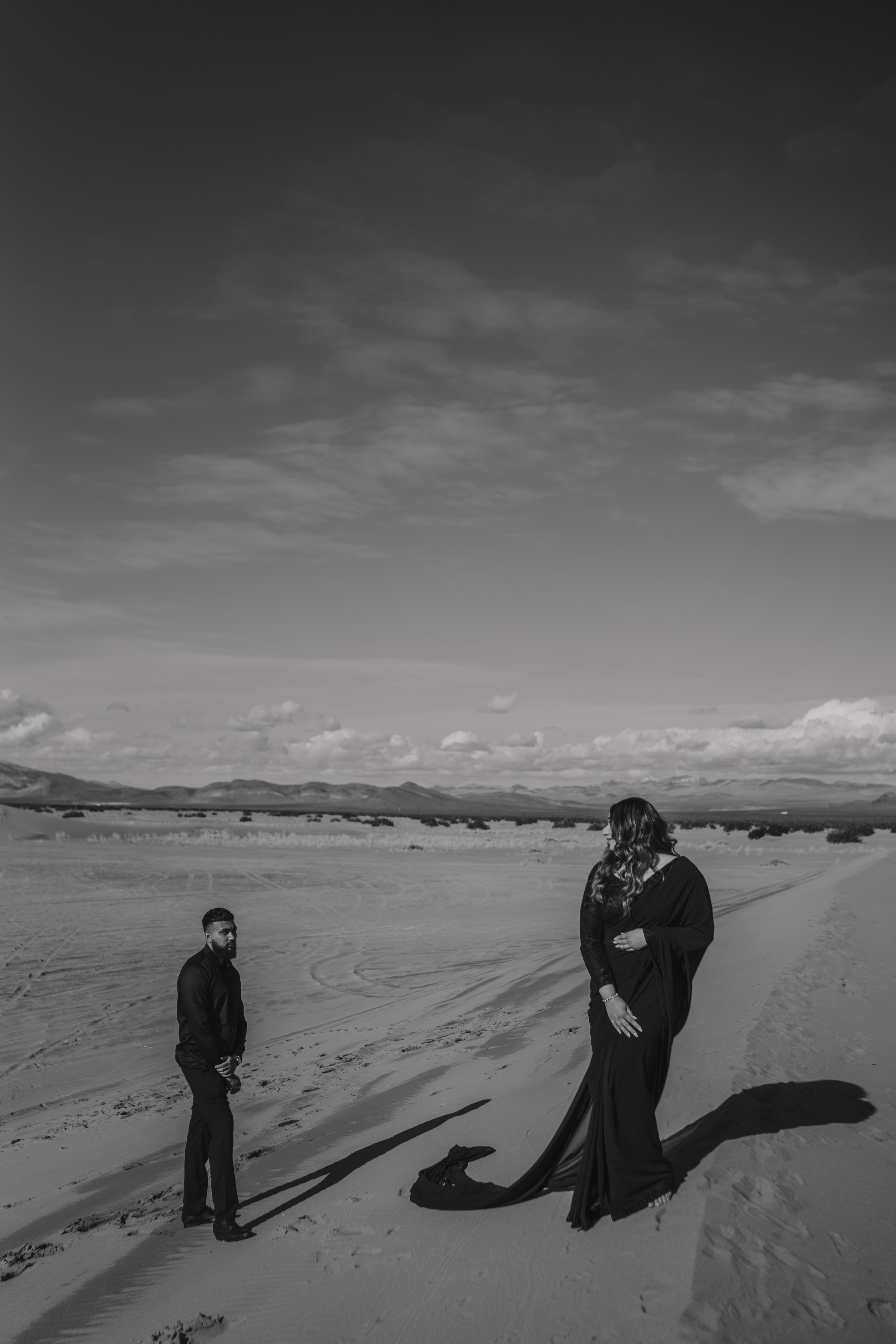 A man and woman standing on a sandy desert landscape with mountains in the distance; the woman is dressed in a long black gown, and the man is in dark clothing, both casting long shadows.