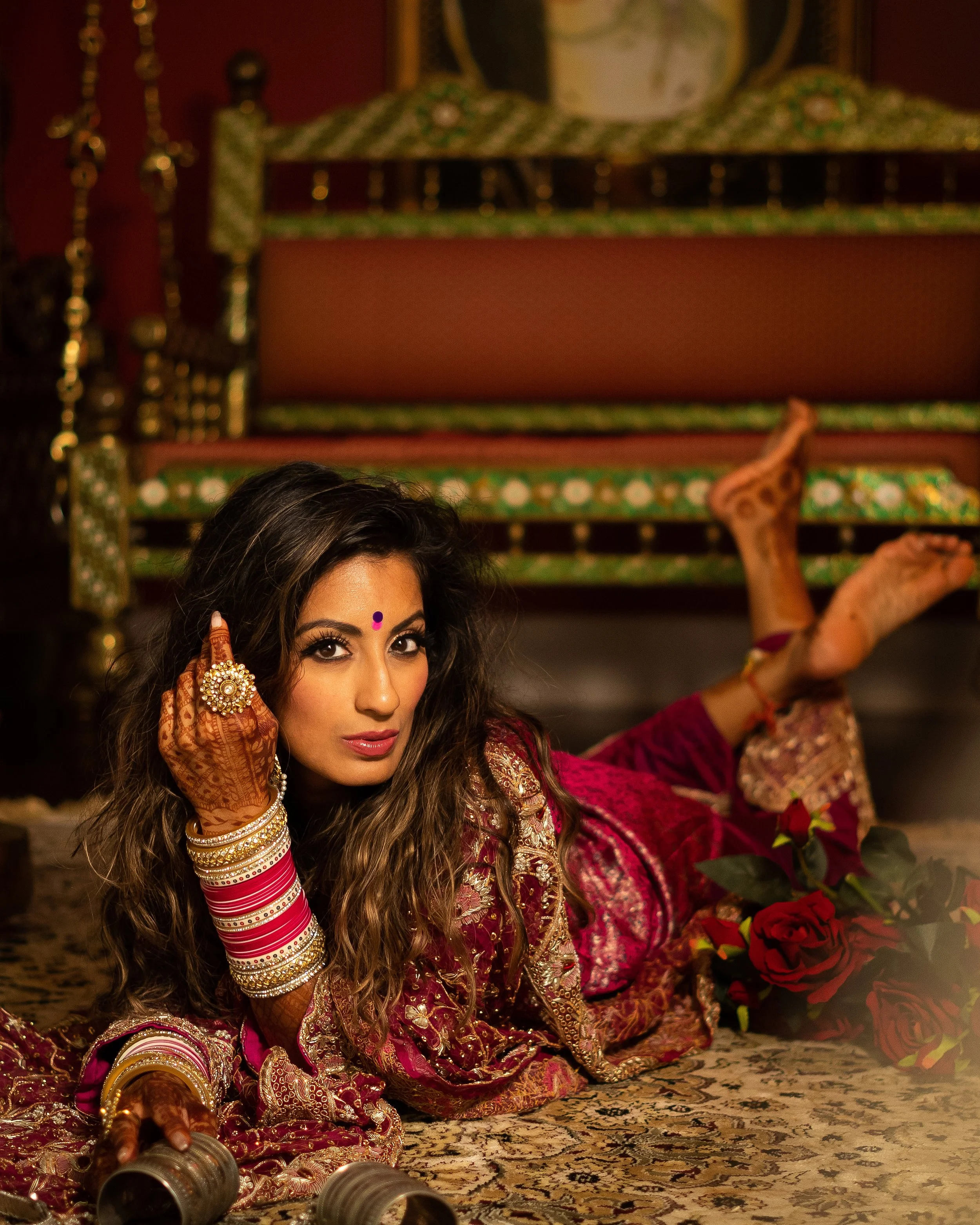 A woman dressed in traditional Indian attire, lying on a carpet, surrounded by red roses, with intricate henna designs on her hands, wearing jewelry, and holding a pair of dance castanets.