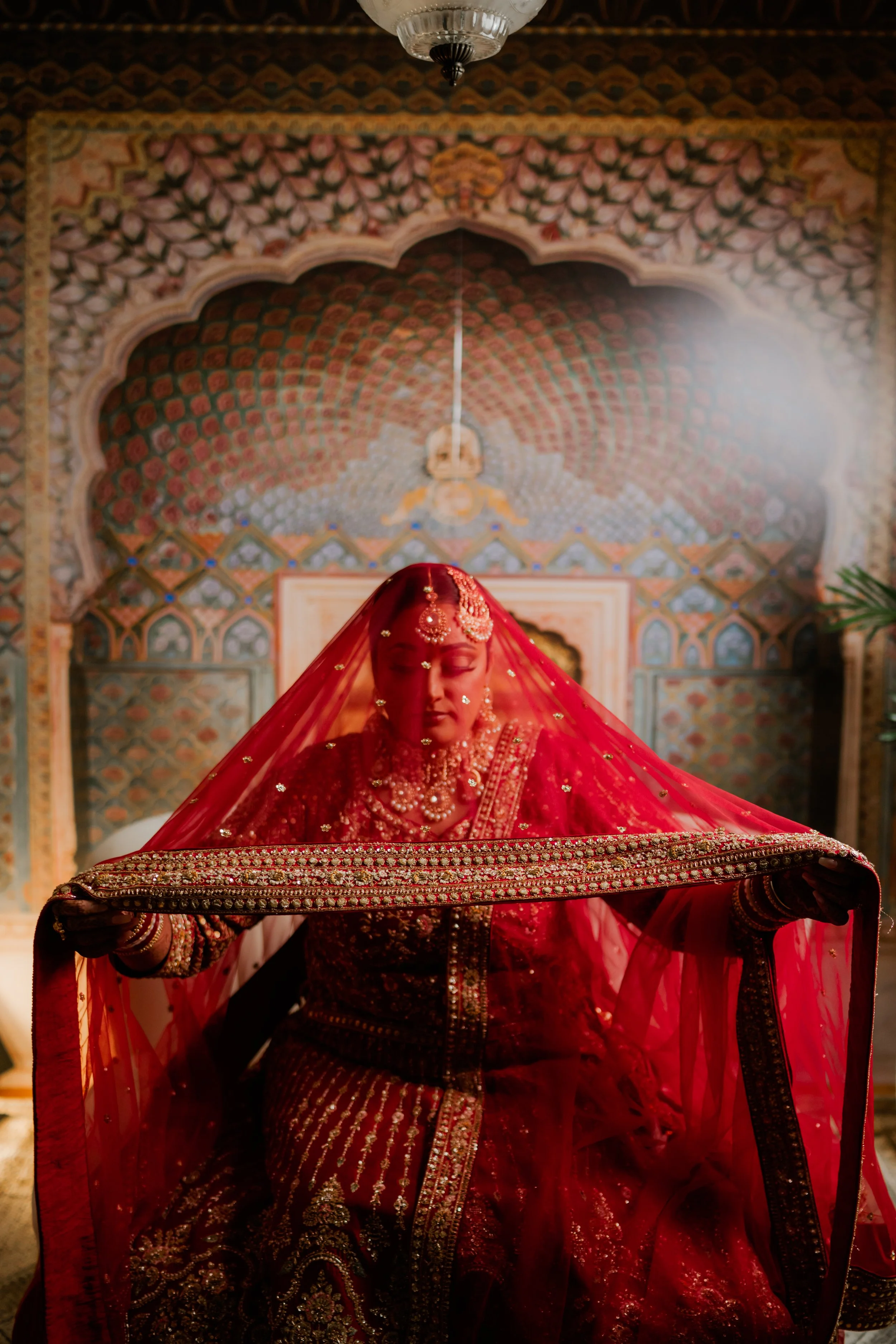 A bride dressed in traditional Indian bridal red and gold attire, sitting with her eyes closed, holding a decorated veil over her head, surrounded by an ornate and colorful backdrop.
