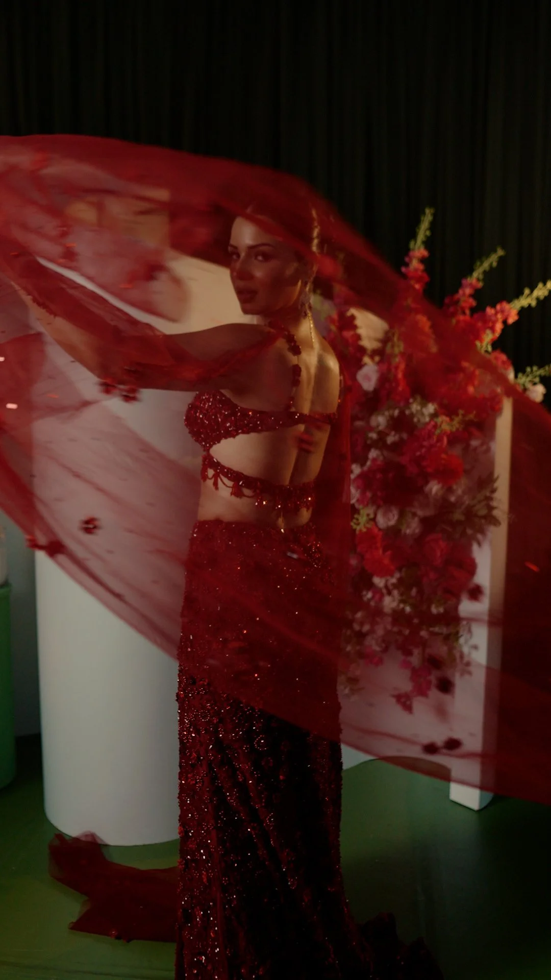 Woman dressed in a red sequined gown with red sheer fabric swirling around her, standing beside a pink and red floral arrangement with black curtains in the background.