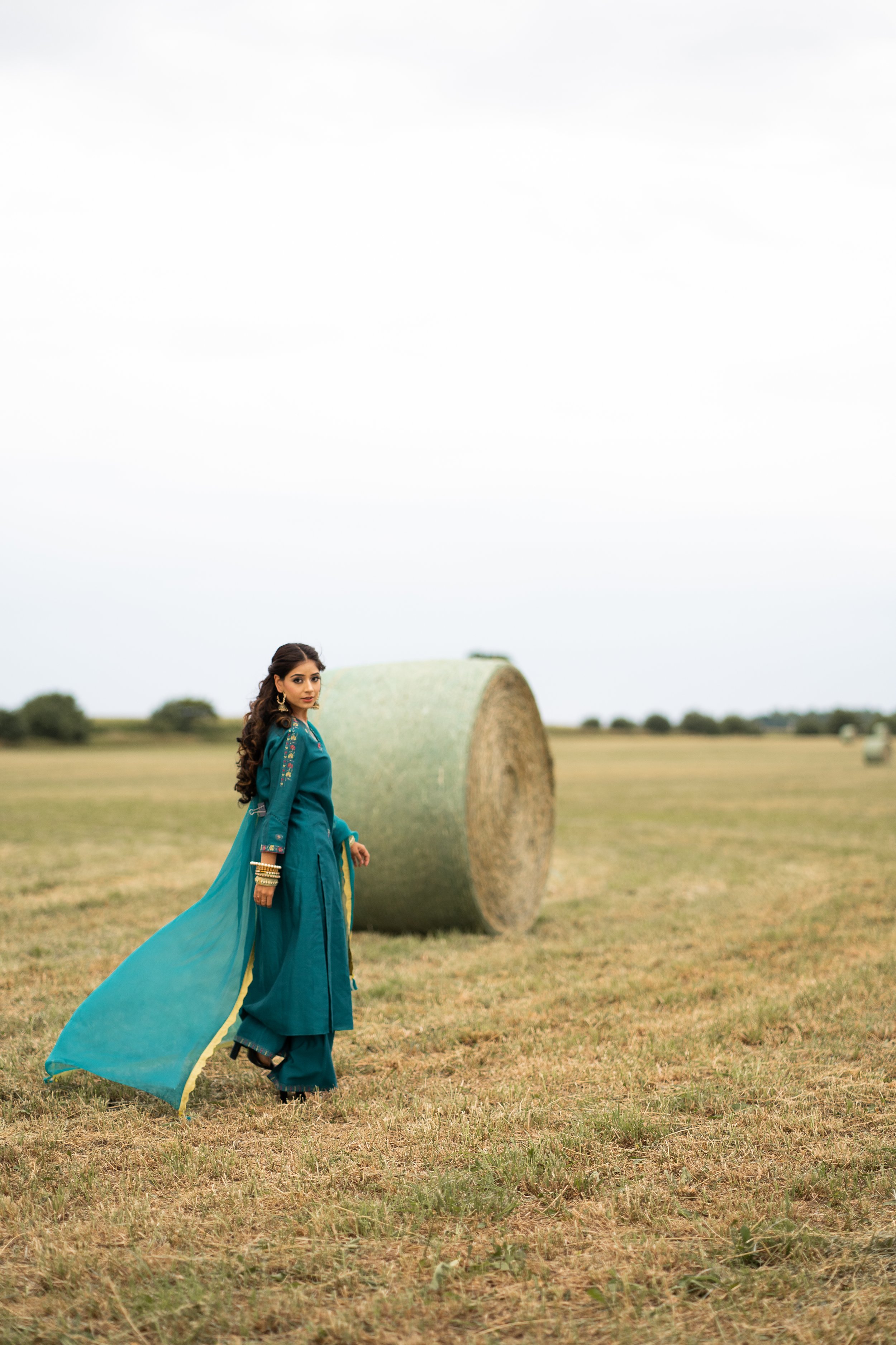 A woman wearing a teal traditional dress with gold embroidery, standing next to a large hay bale in an open field during daytime.