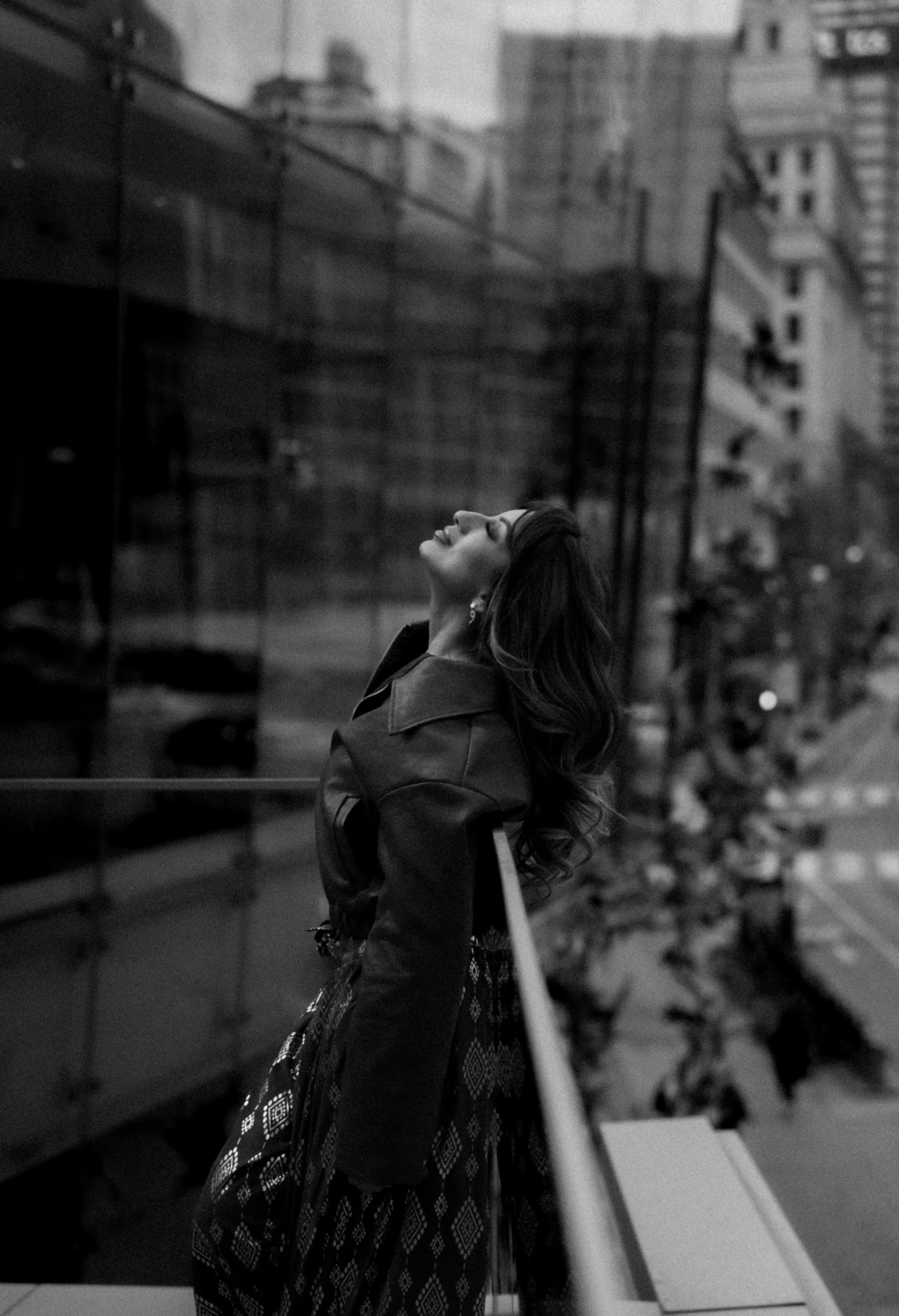 A woman with long wavy hair standing on a balcony with her head tilted back and eyes closed, wearing a dark jacket and patterned skirt, in an urban city setting with buildings in the background.
