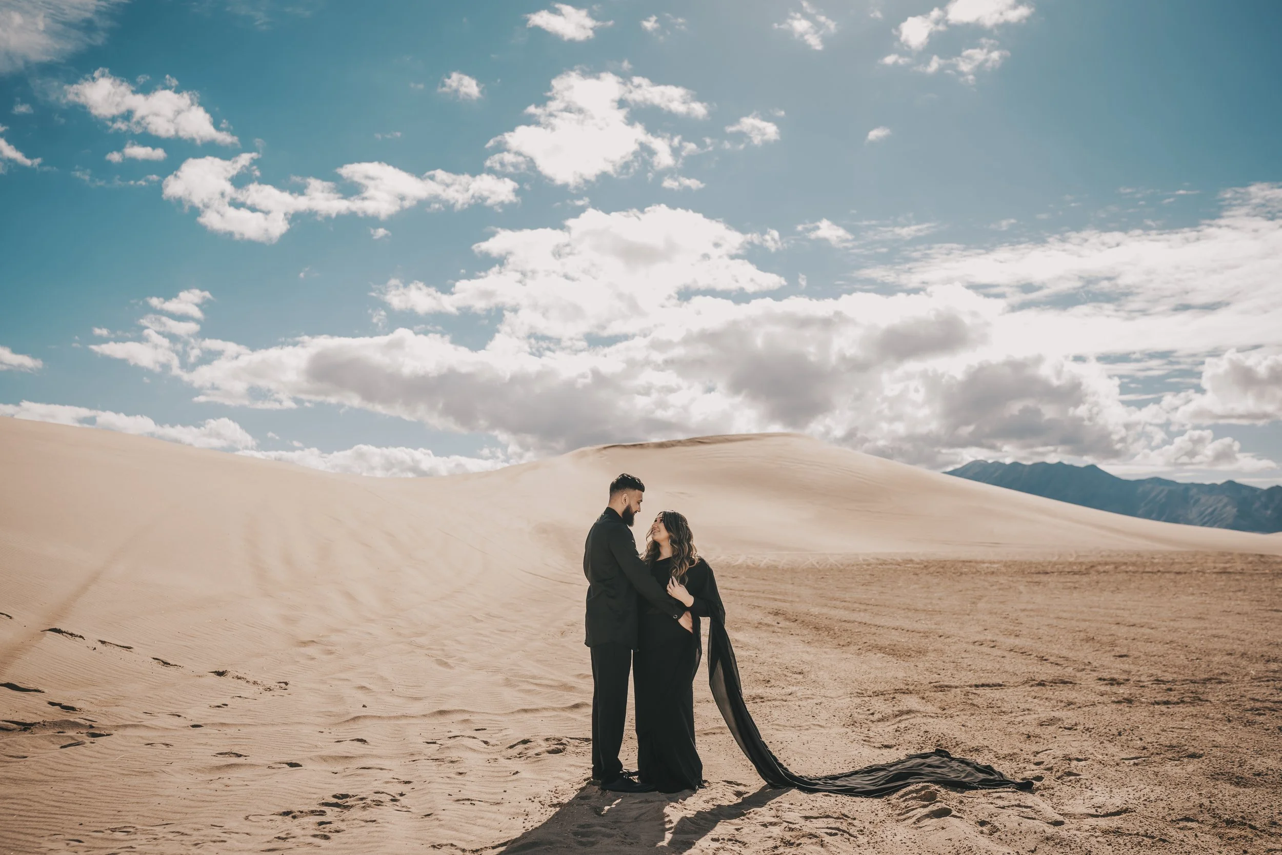 A couple dressed in black standing close together in a desert landscape with sand dunes, blue sky, and scattered clouds.