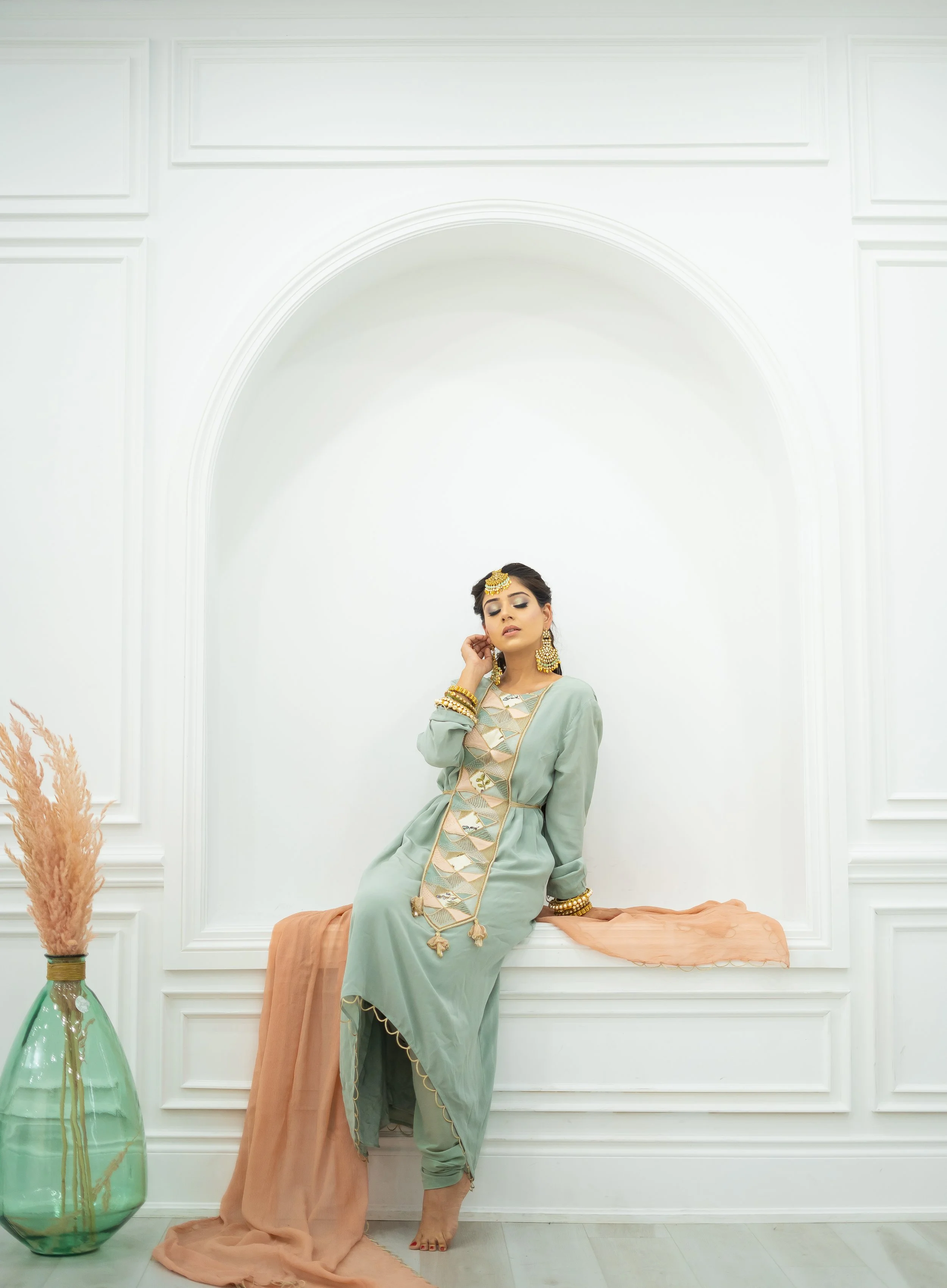 A woman dressed in traditional attire with jewelry, sitting on a window ledge in a white room, next to a green vase with dried pampas grass.