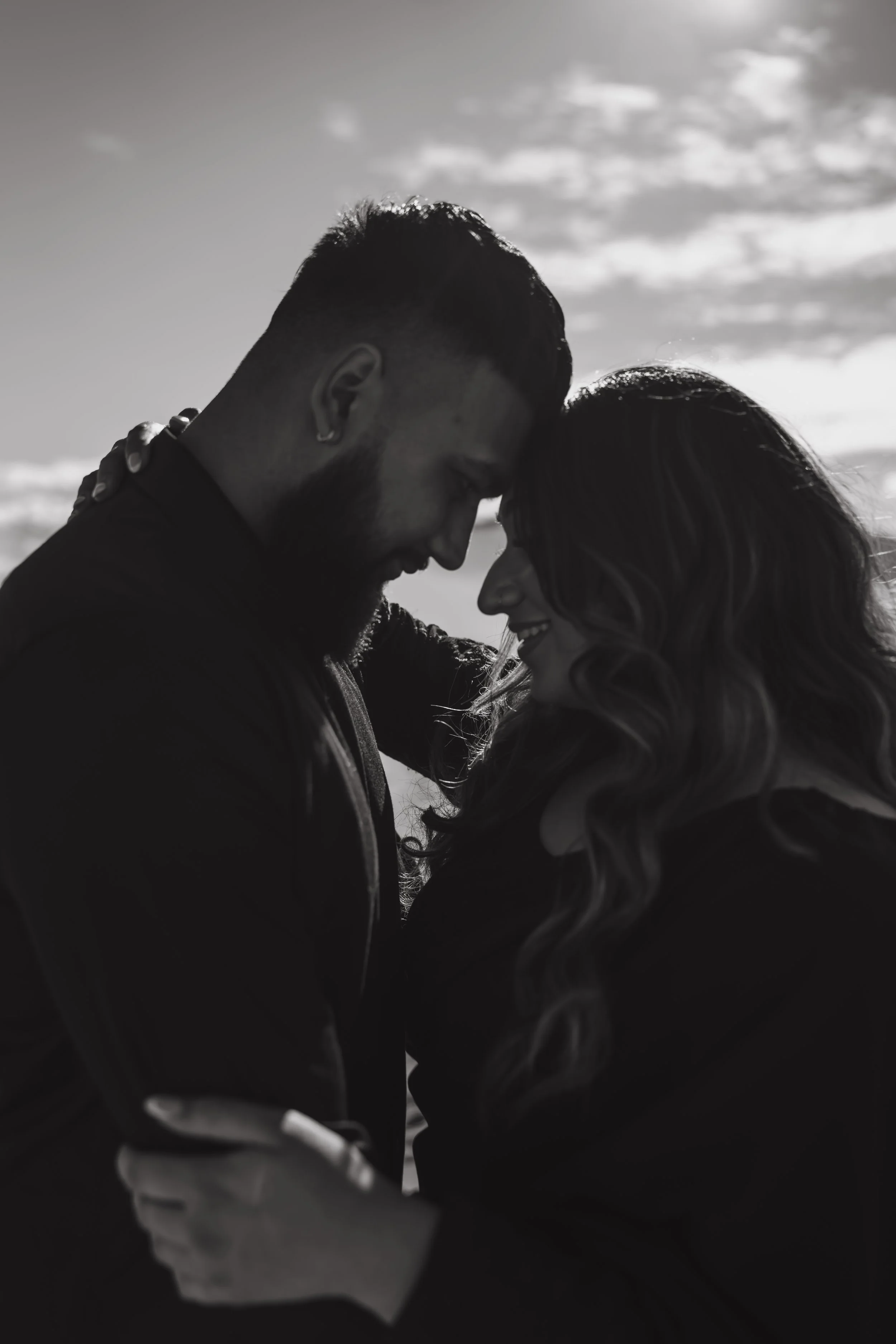 A black and white photo of a couple with their foreheads touching, smiling, and holding each other during a moment of intimacy outdoors with a cloudy sky in the background.