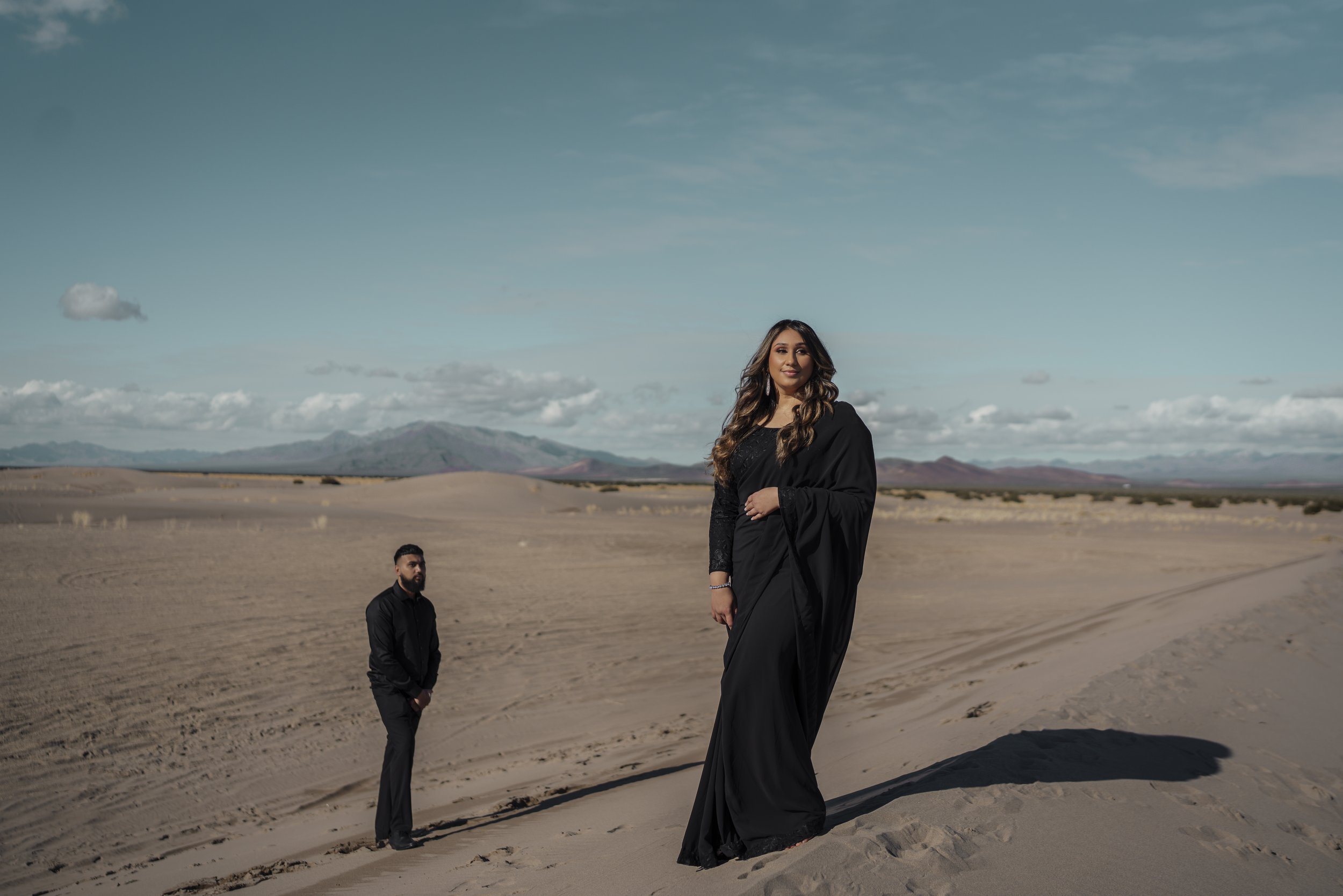 A woman in a black dress standing on a sand dune in a desert, with a man in black clothing standing in the background, under a blue sky with scattered clouds.