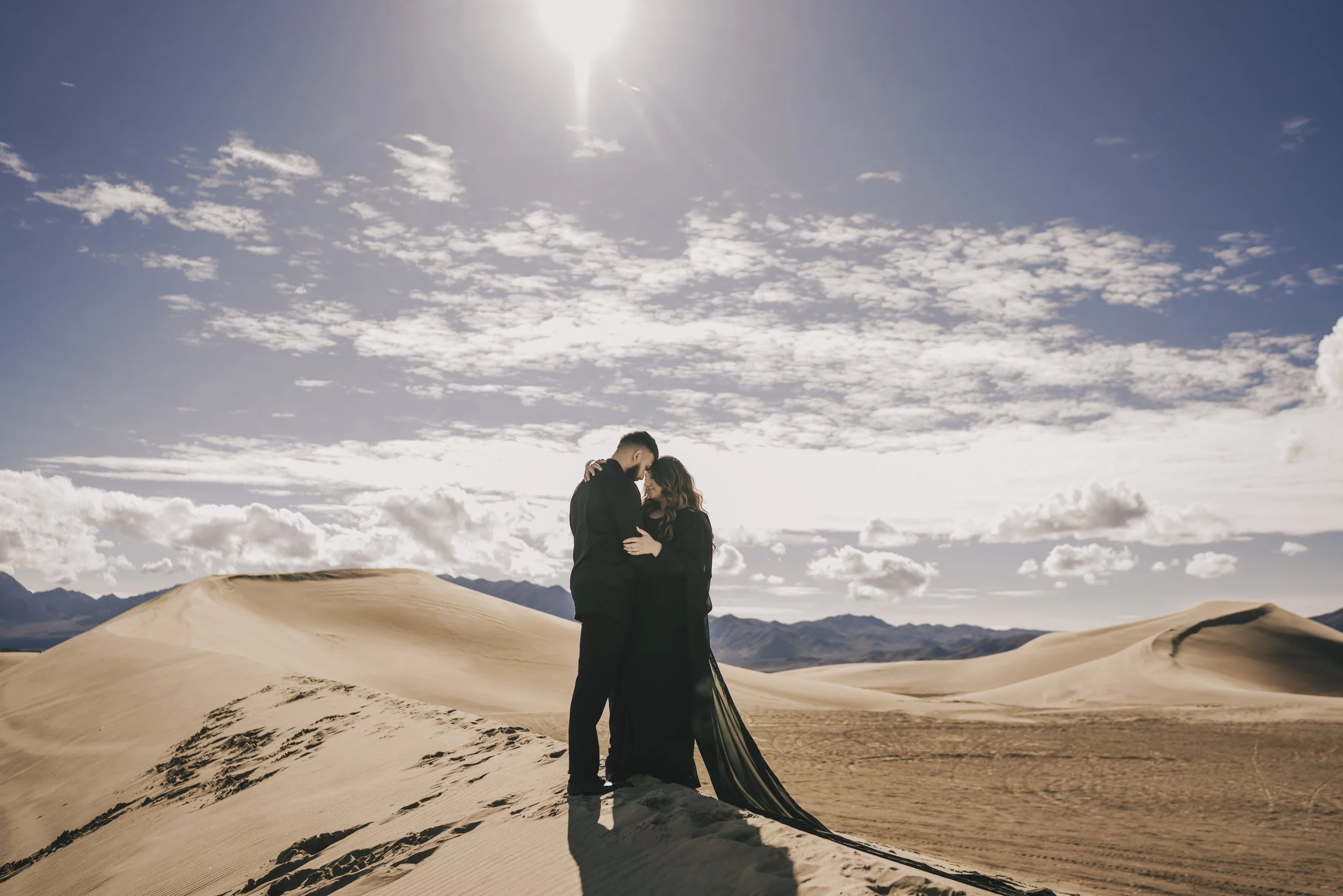 A couple in black clothing embracing in a desert with sand dunes and mountains in the background under a partly cloudy sky with sunlight.