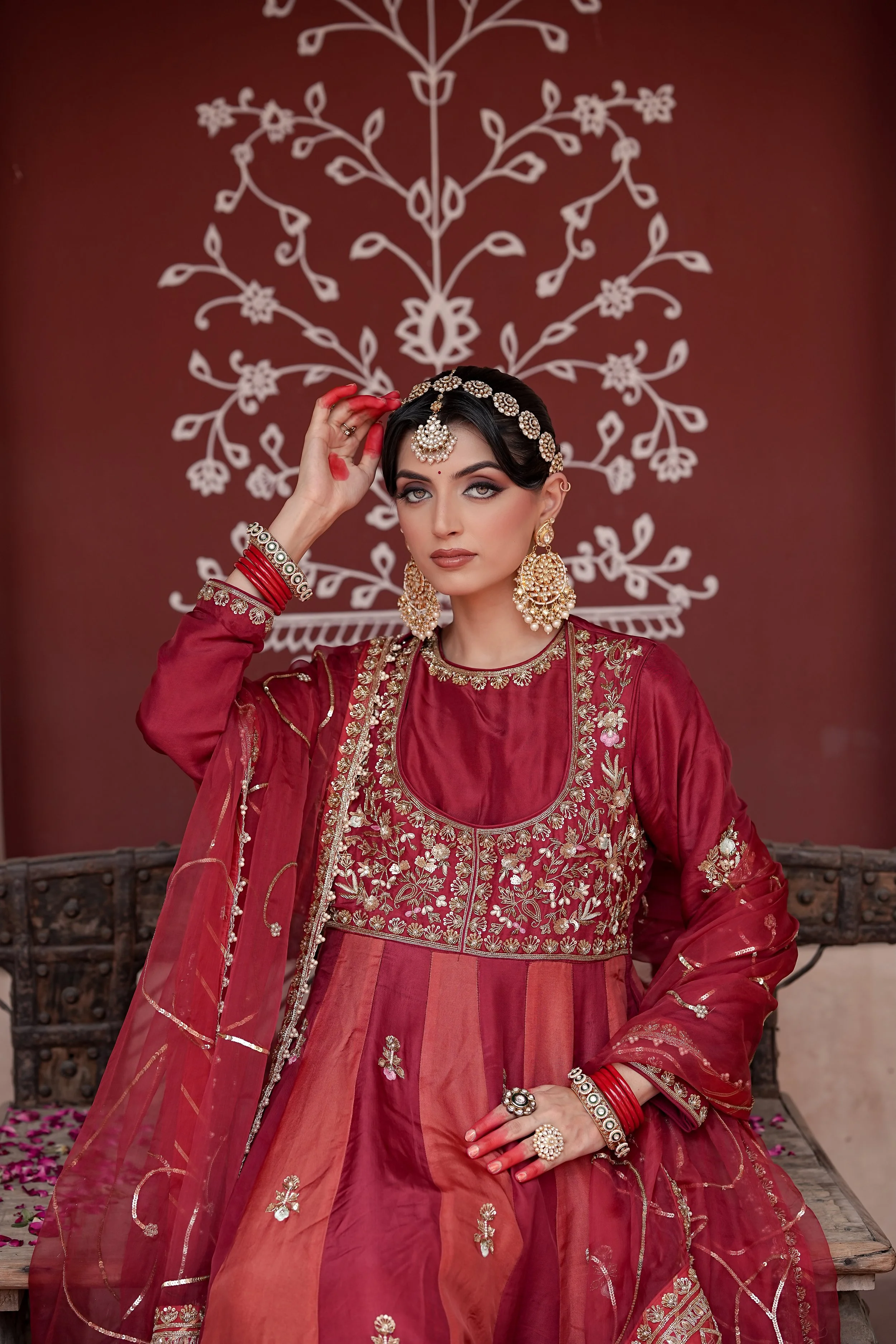 A woman dressed in traditional Indian attire, including a red embroidered dress and heavy gold jewelry, standing against a backdrop with a white floral design.