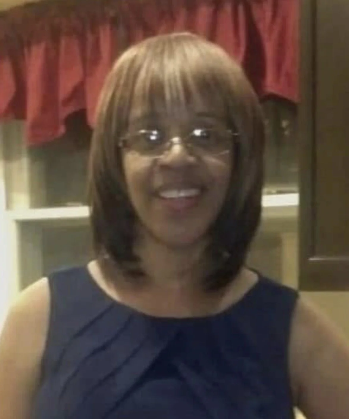 A woman smiling, wearing glasses and a navy blue dress, with red curtains in the background.