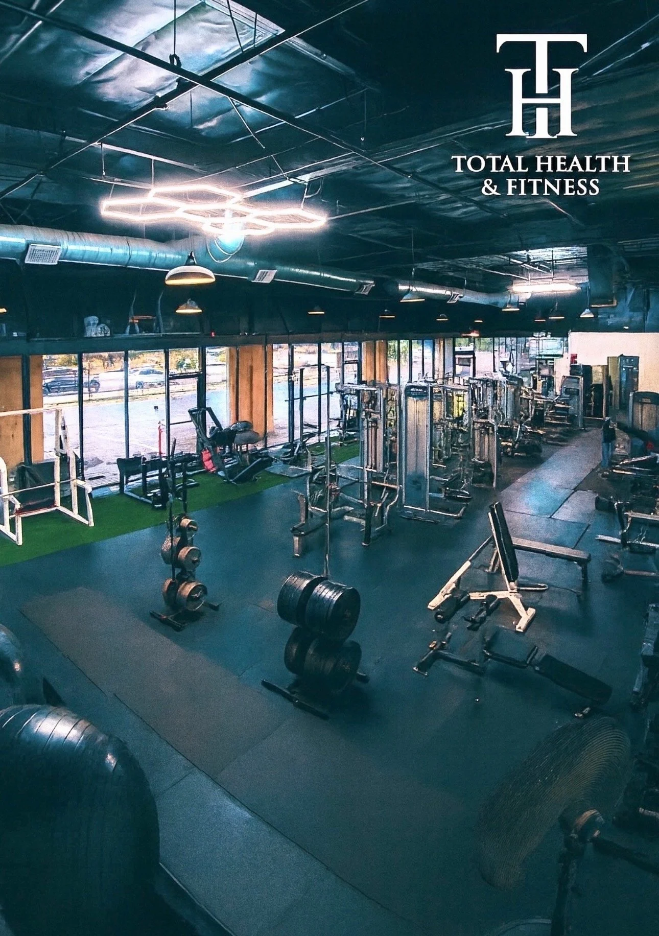 Interior of a gym with various exercise equipment, including weights, machines, and cardio equipment, with large windows and a black ceiling.