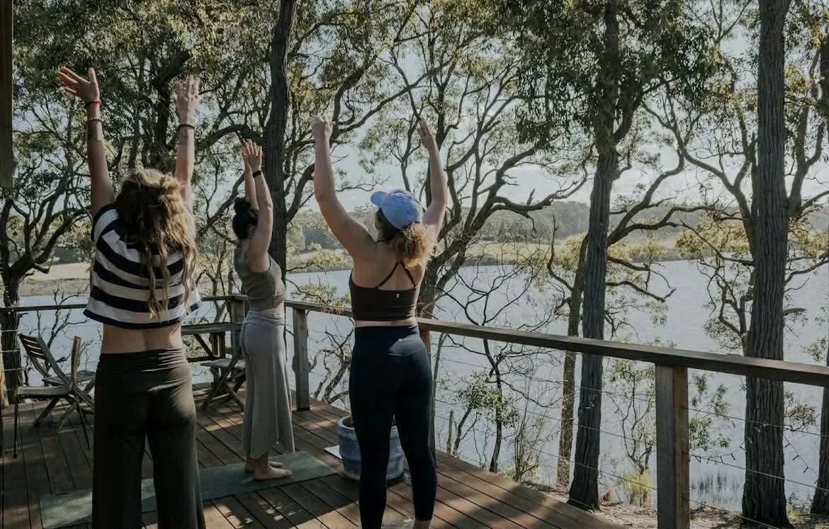 Three women practicing yoga on a wooden deck overlooking a lake surrounded by trees, with their arms raised in a yoga pose.