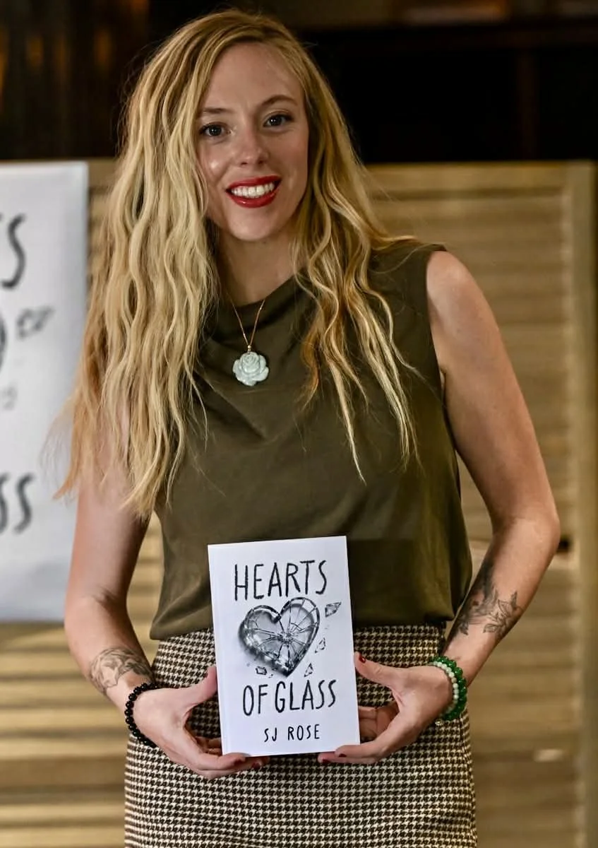 A woman with long blonde curly hair wearing a sleeveless dark green top, a necklace with a flower pendant, and patterned skirt, holding a book titled 'Hearts of Glass' by SJ Rose, smiling at the camera.