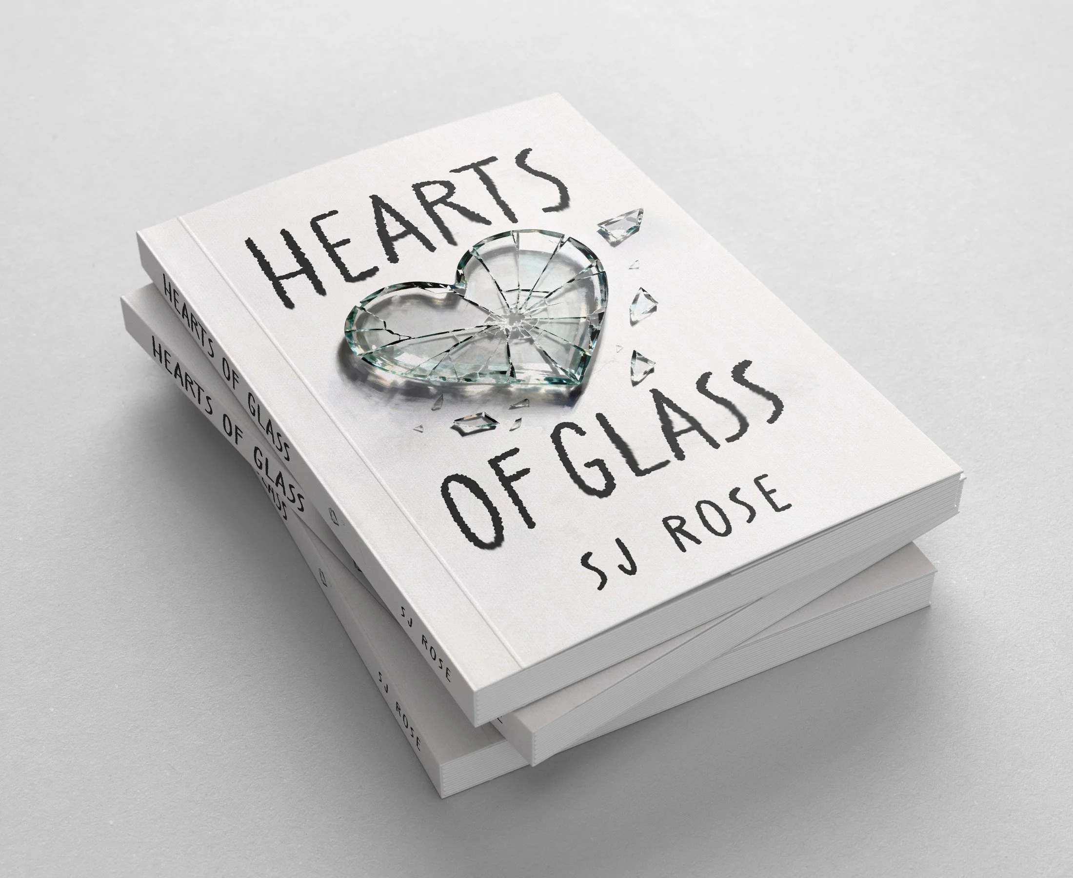 Three stacked white books with the title "Hearts of Glass" by SJ Rose, with a cracked glass heart on top, some shards scattered around, all on a gray surface.