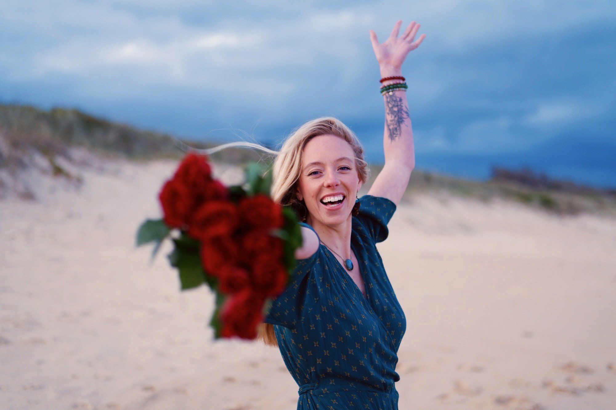 Woman on a sandy beach holding a bouquet of red flowers, smiling, with her arm raised, blonde hair, wearing a blue dress, with a tattoo on her arm, cloudy sky in the background.