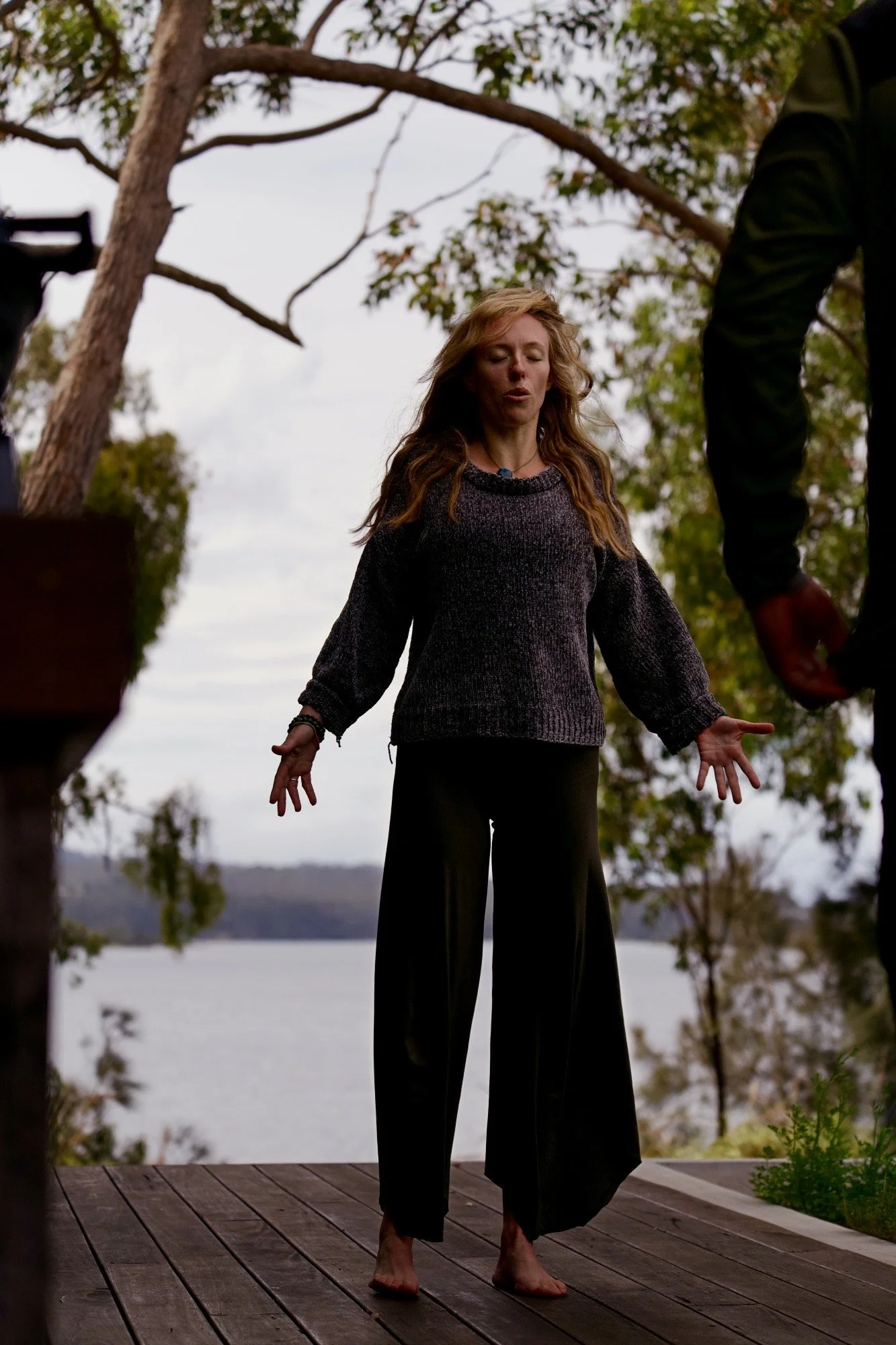 A woman with long, wavy hair standing barefoot on a wooden dock outdoors, with trees and a body of water in the background, during daytime.