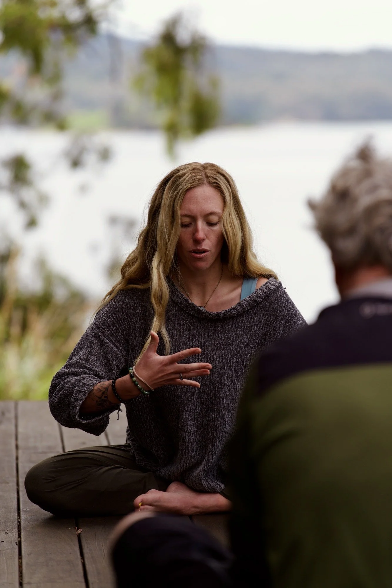 Woman with long wavy blonde hair sitting cross-legged outdoors, speaking and gesturing with her hand, with lake and trees in the background.