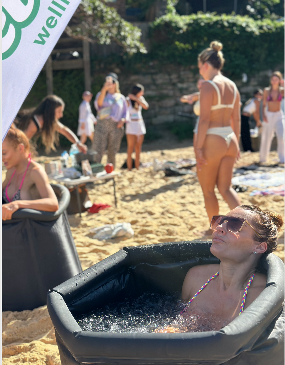 A woman wearing sunglasses and a colorful swimsuit top relaxes in a black inflatable tub on a sandy beach. In the background, several women are standing, sitting, or walking on the sand, some taking photos and others chatting, with greenery and rocks