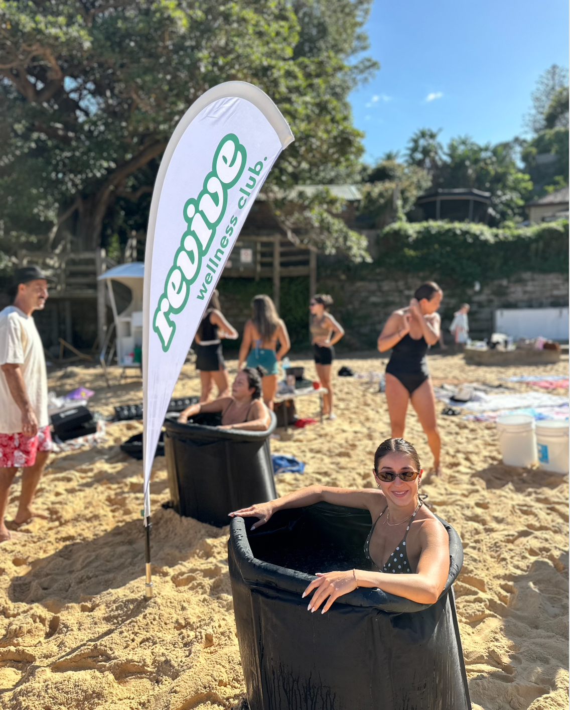 People participating in a wellness activity at the beach, with a banner reading 'reboot wellness club' visible in the foreground.
