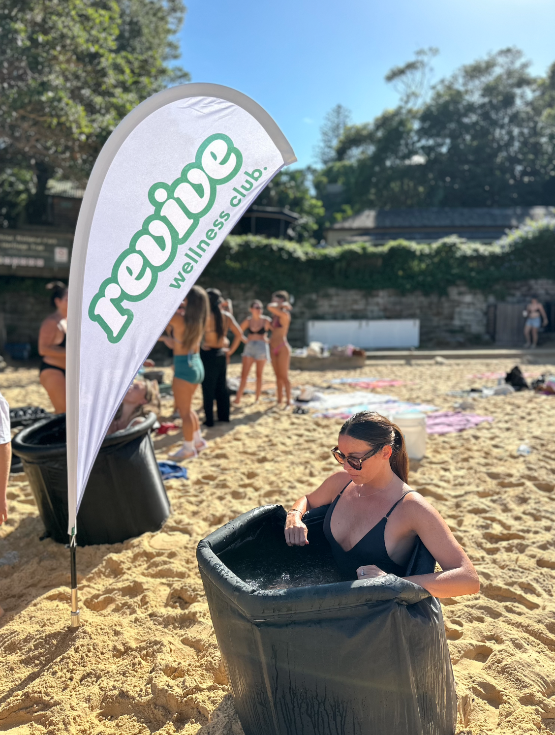 Five women practicing yoga in a tree pose on a beach with ocean waves and foggy hills in the background.