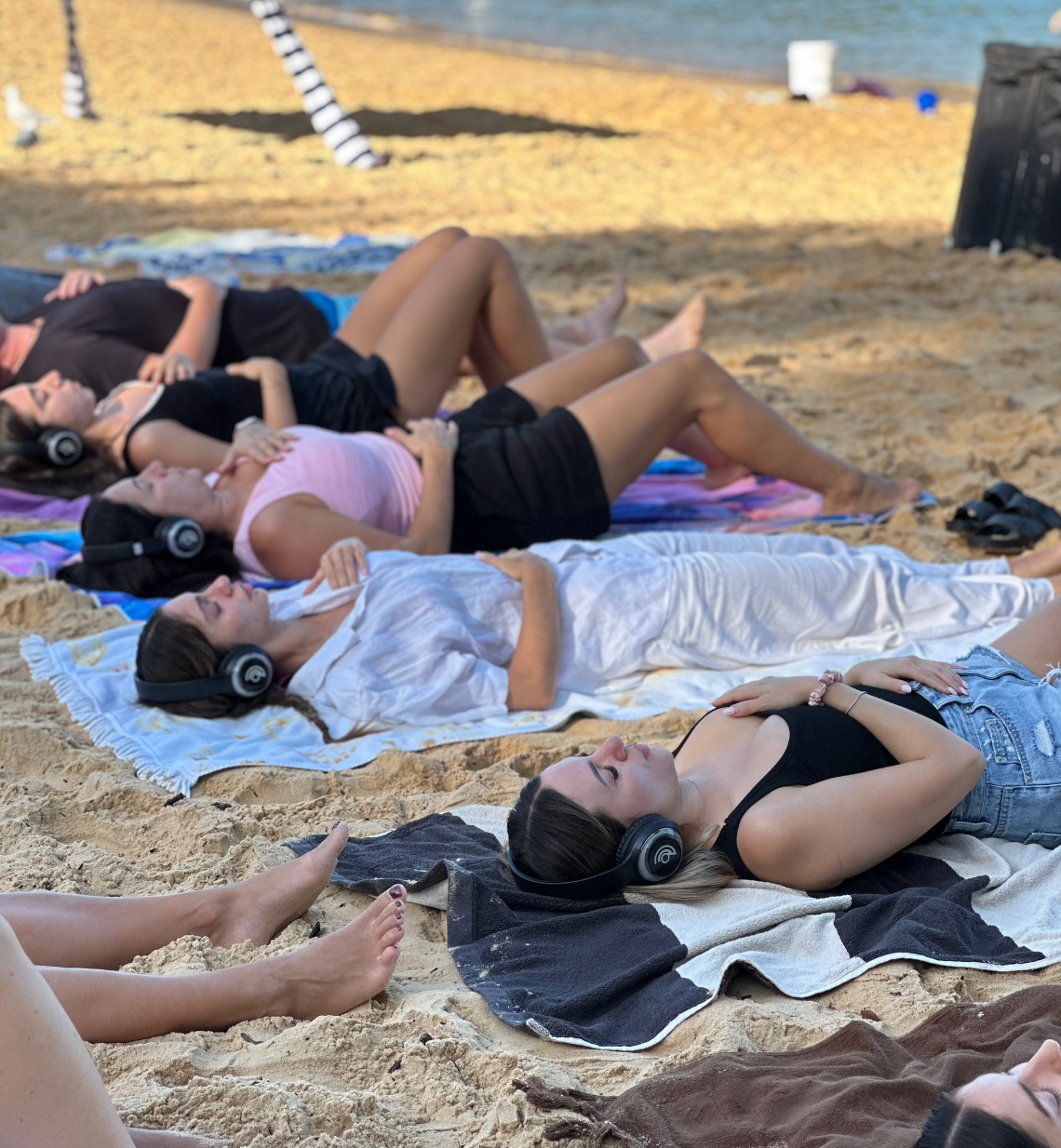 Group of people doing 9D Breathwork with headphones on the beach, lying on towels in a line near the ocean.