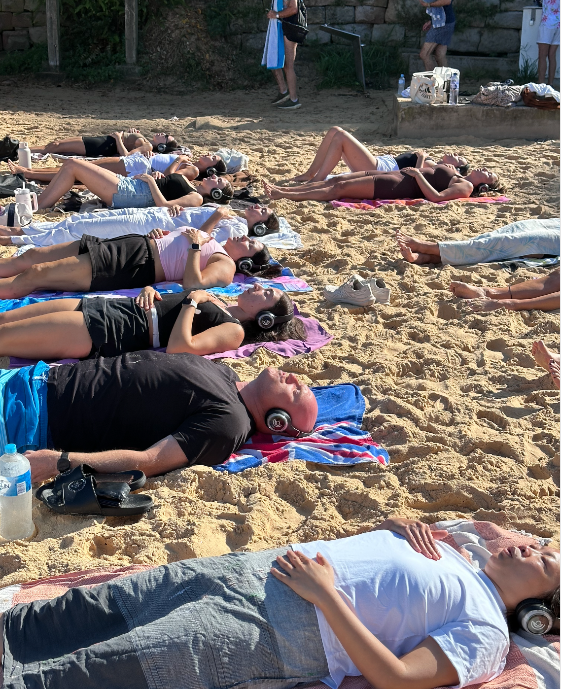 Two people relaxing outdoors on yoga mats by a pool, wearing eye masks and headphones, sunbathing on a sunny day.