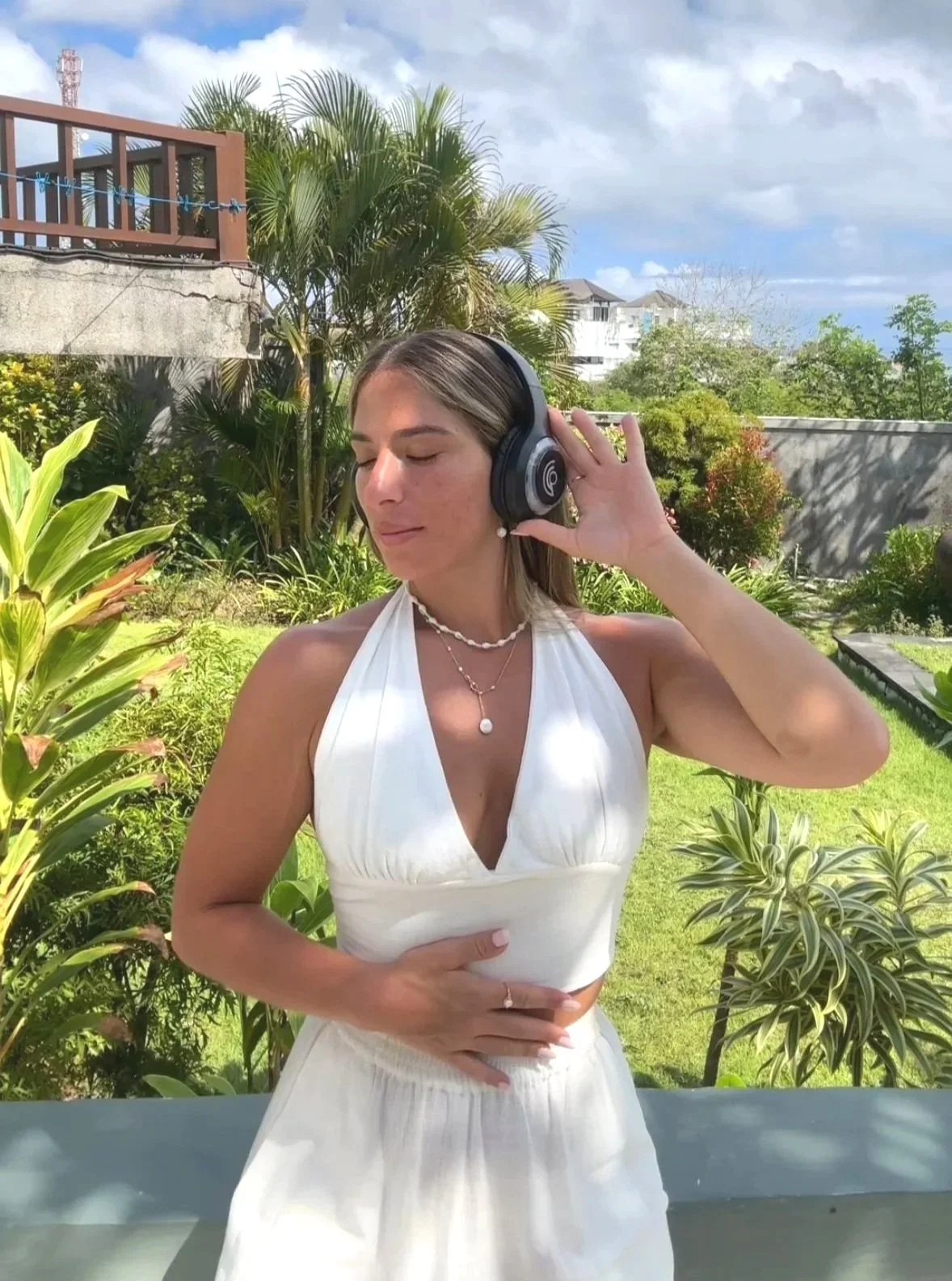 A woman with light skin and brown hair is outdoors in a garden, wearing a white halter top and white skirt. She is listening to music with large black headphones, eyes closed, with one hand on the headphones and the other on her stomach. There are green plants and trees in the background, with a partly cloudy sky.