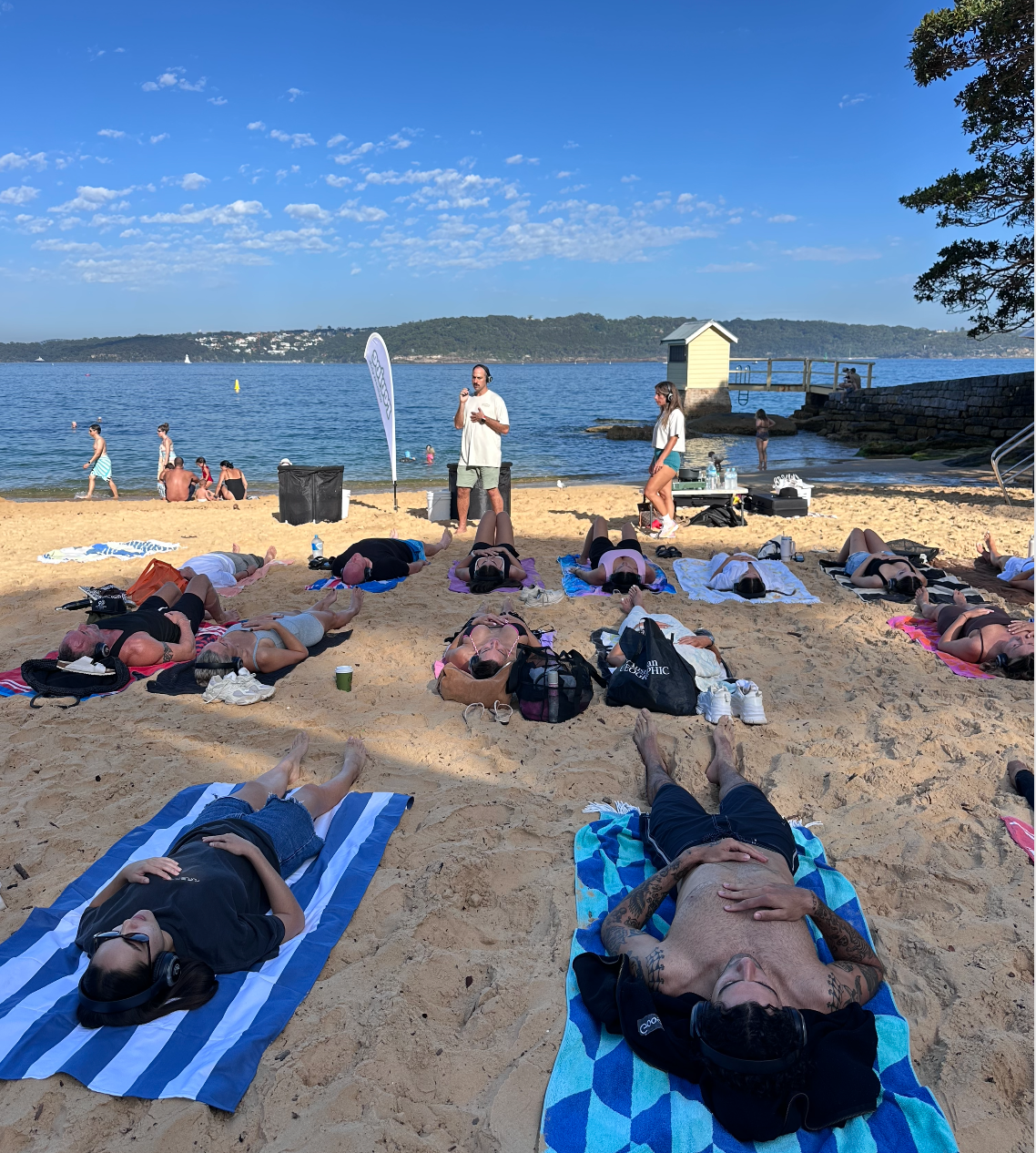 People relaxing and participating in a yoga or meditation session on a sandy beach near the water, with a clear blue sky, some swimmers and a small yellow building in the background.